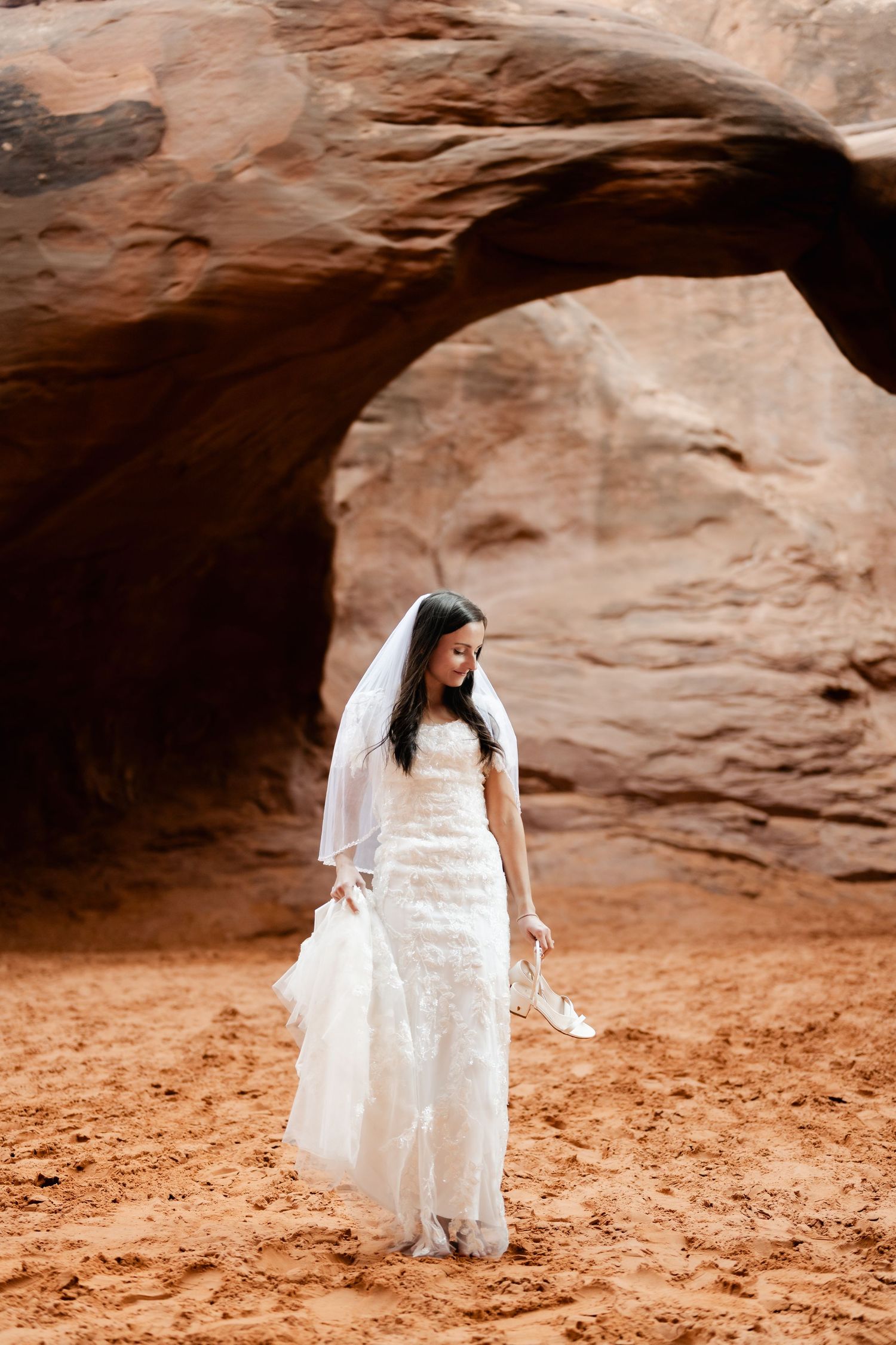 Bride in lace wedding dress standing under a natural rock arch in the desert.