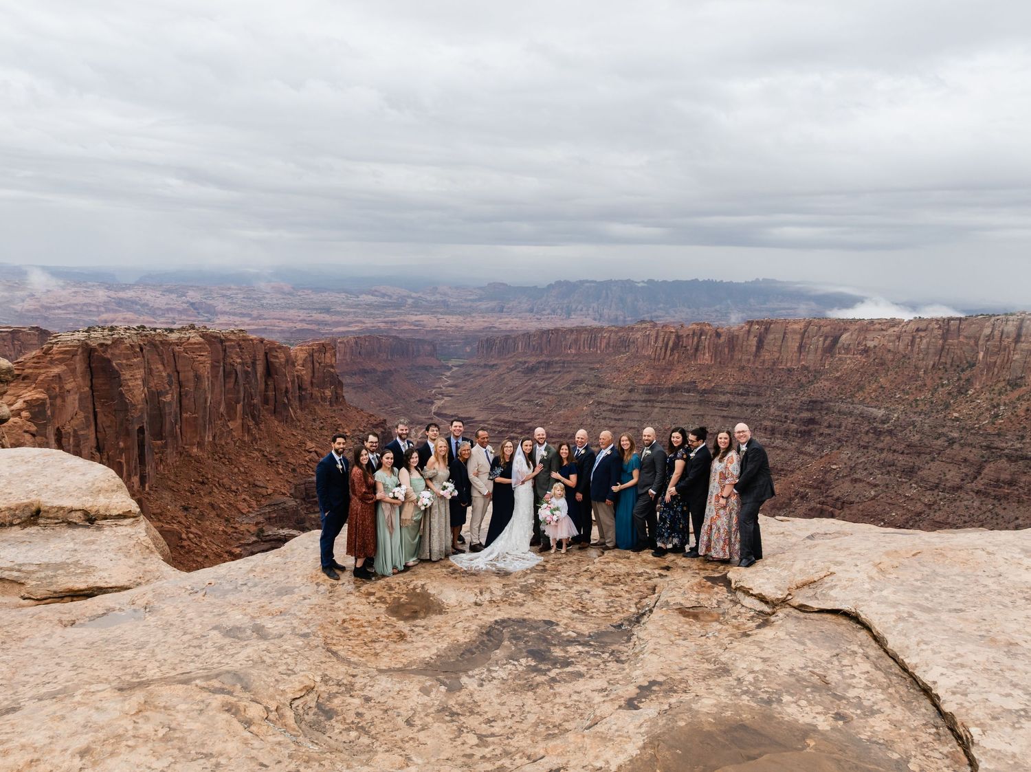 A wedding party poses on the edge of a dramatic canyon overlook with red rock cliffs and cloudy skies in the background.