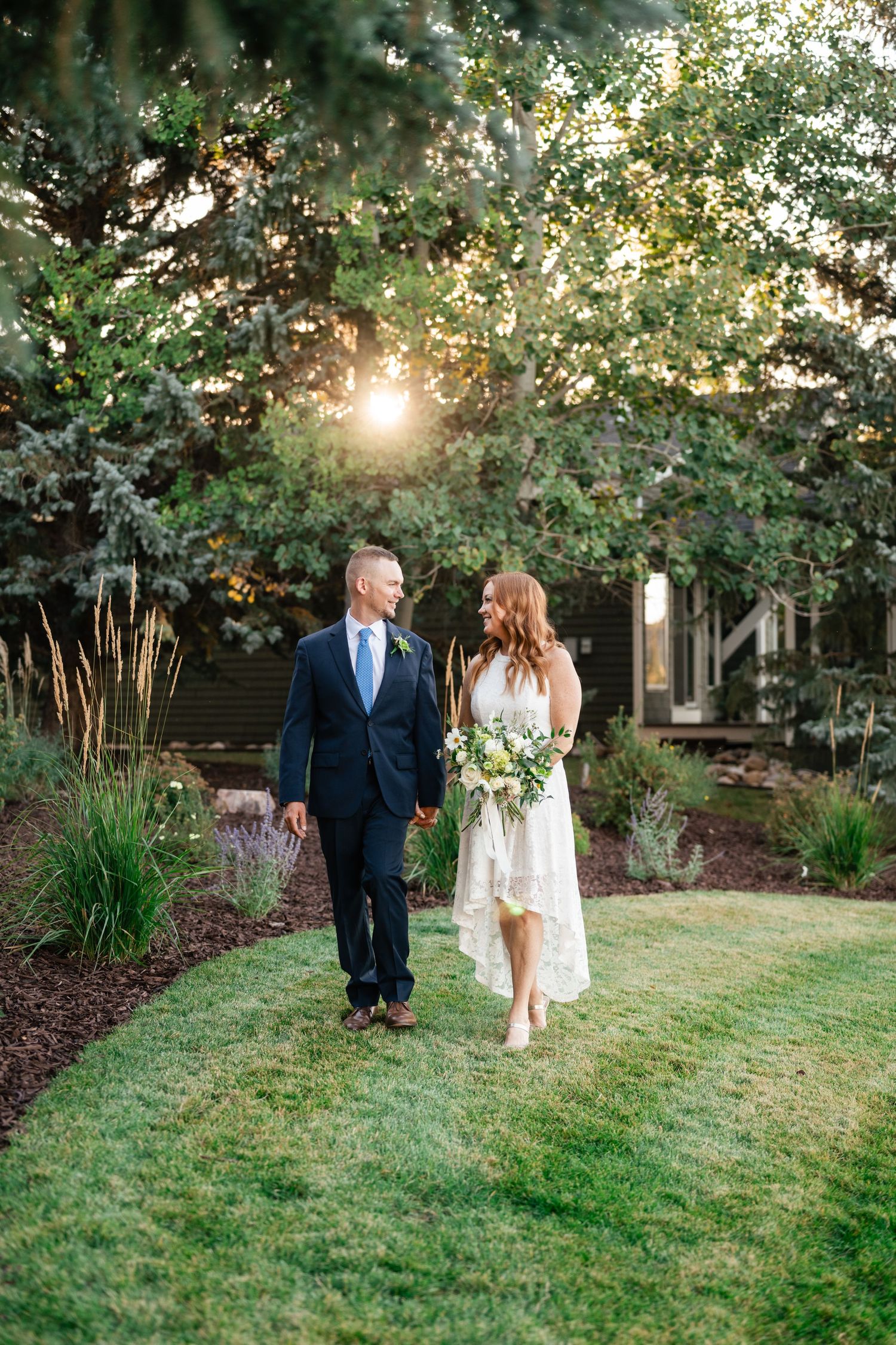 A couple walks together on a lush green lawn surrounded by trees during golden hour.
