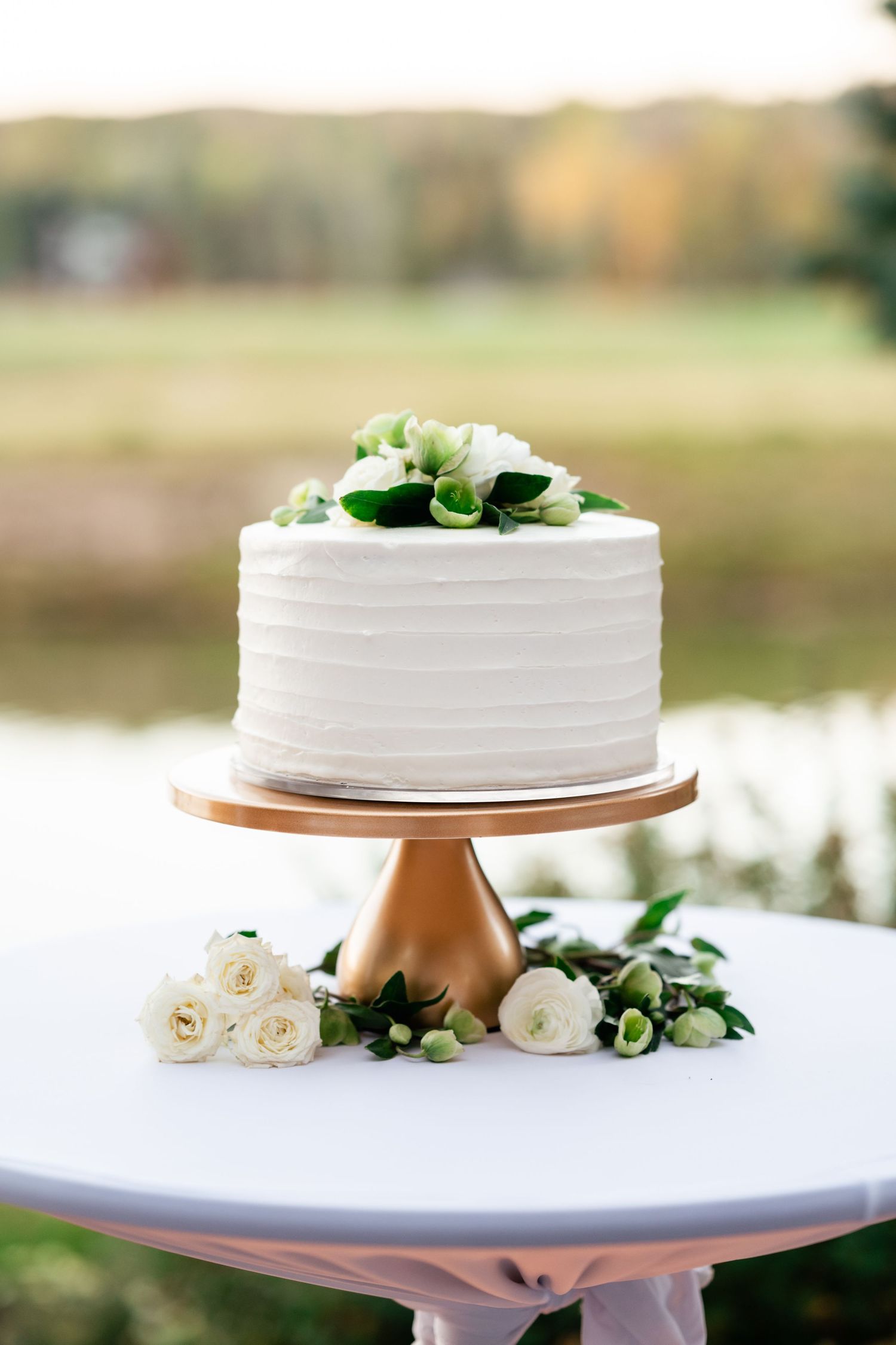 White wedding cake decorated with fresh flowers displayed on a stand outdoors.