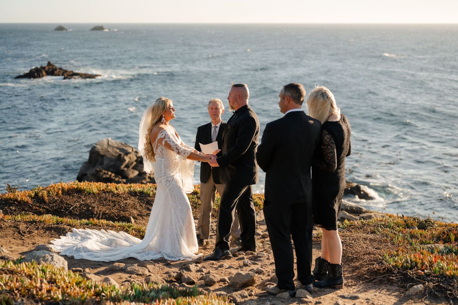 Wedding party stands together on rocky coastal cliffs with ocean views at sunset.