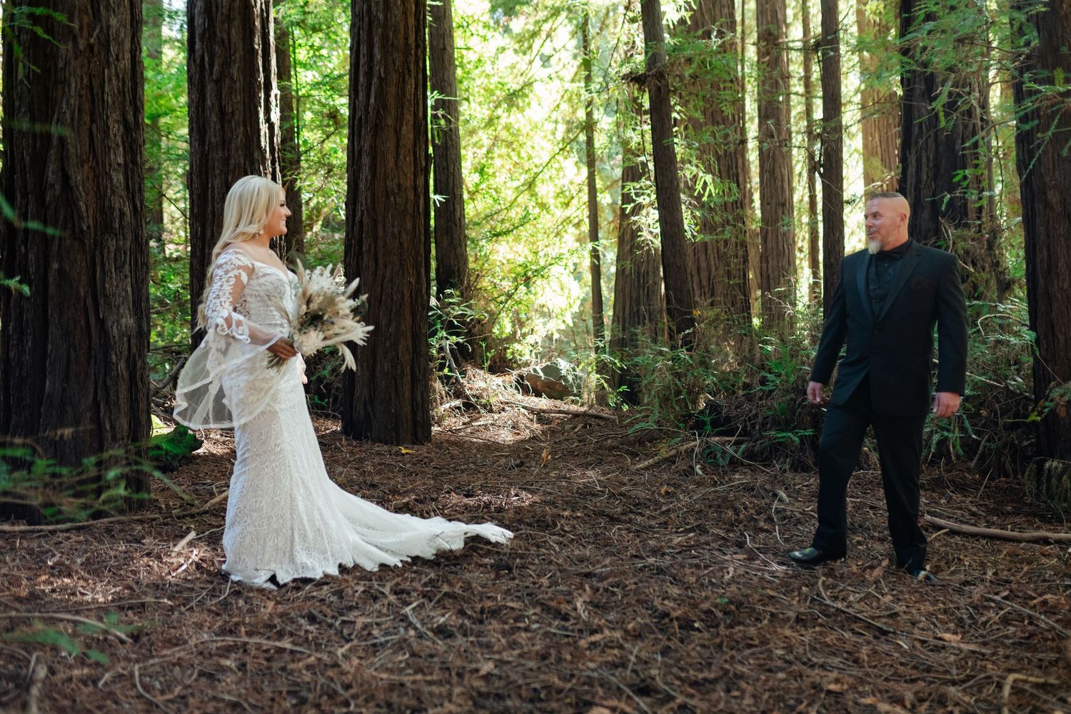 A wedding couple shares a first look moment among towering redwood trees in a forest setting.
