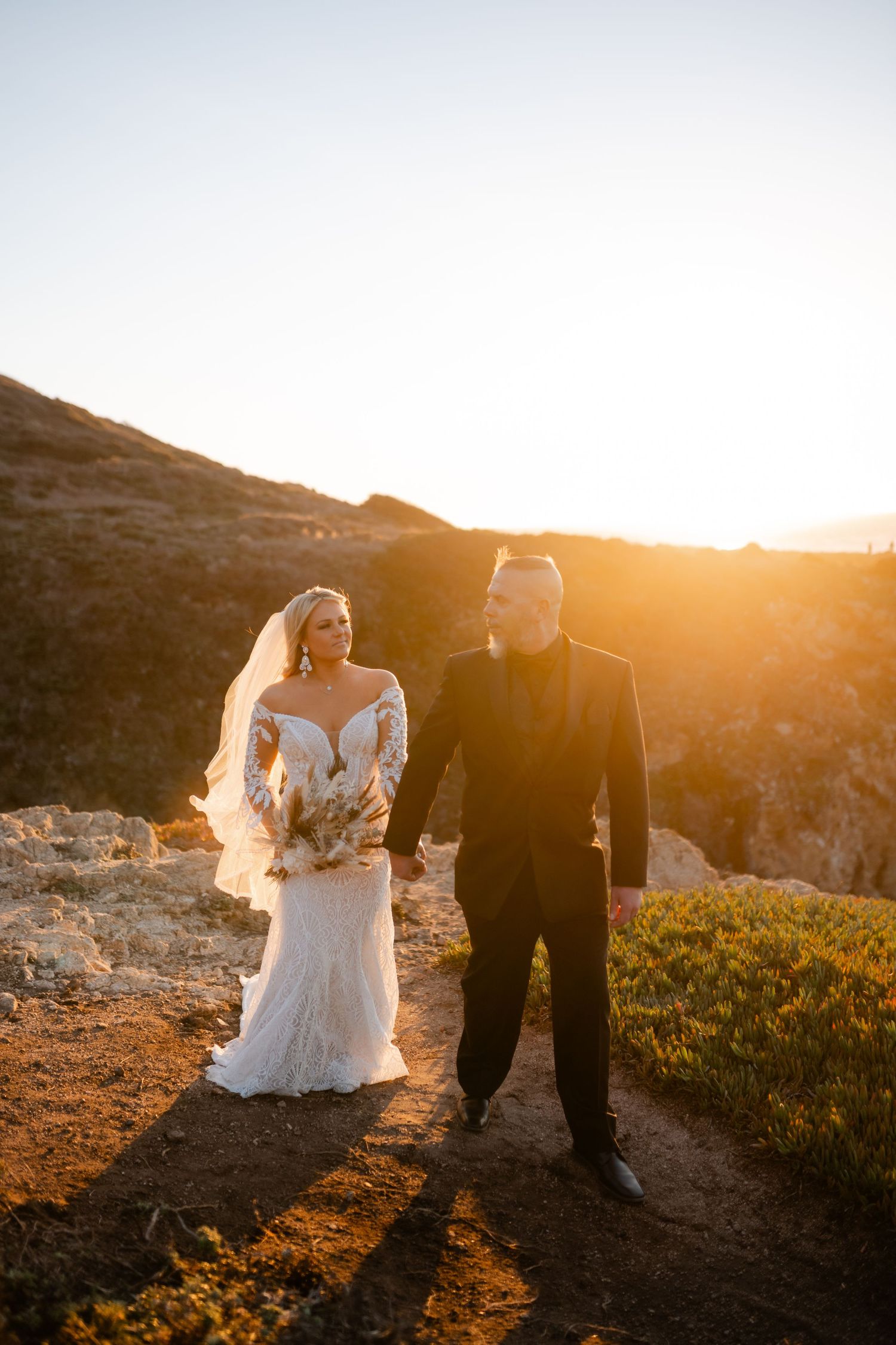 Couple walks along scenic coastal path during golden hour sunset wedding photoshoot.