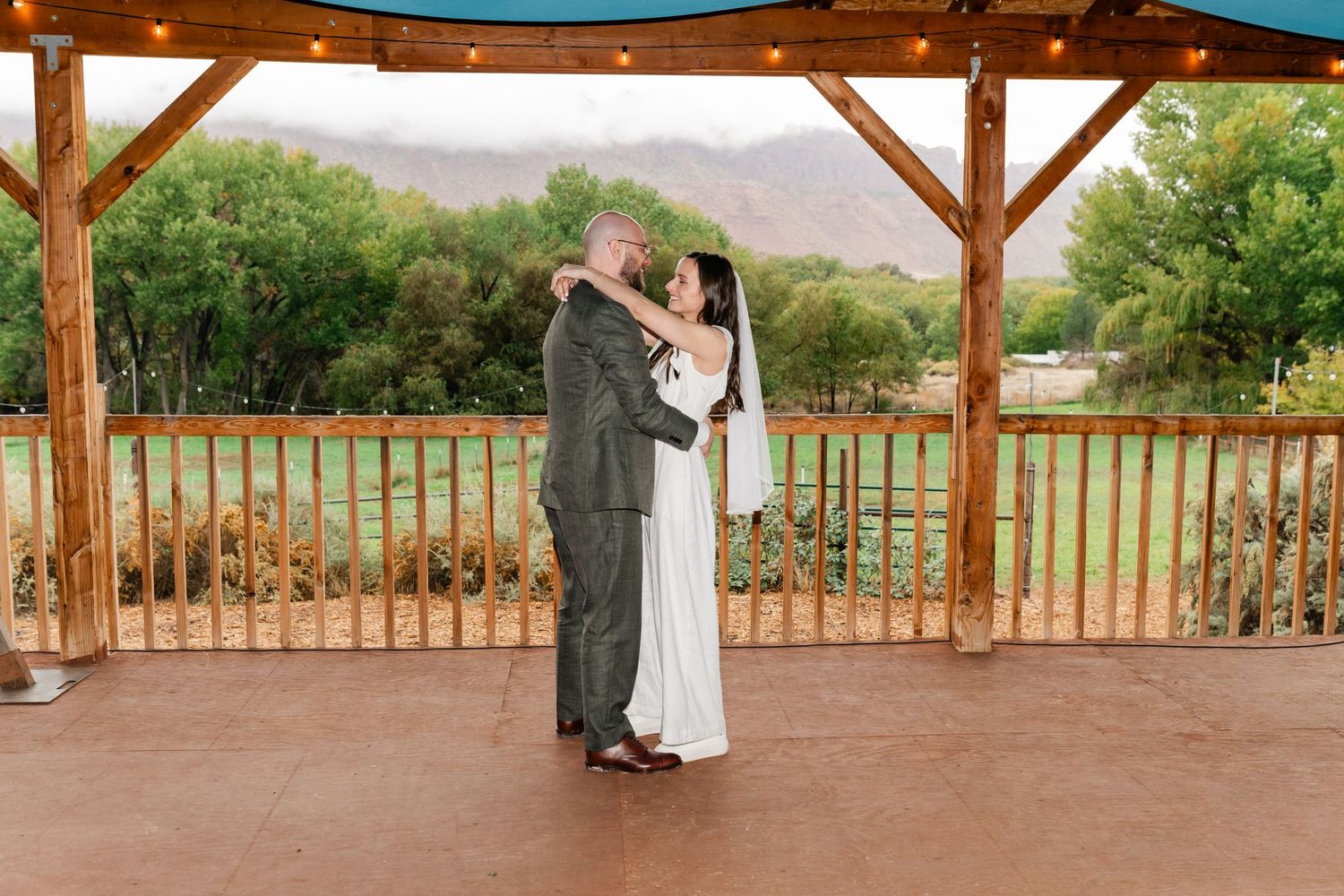 Wedding couple dances on an outdoor covered patio with scenic mountain backdrop and string lights overhead.