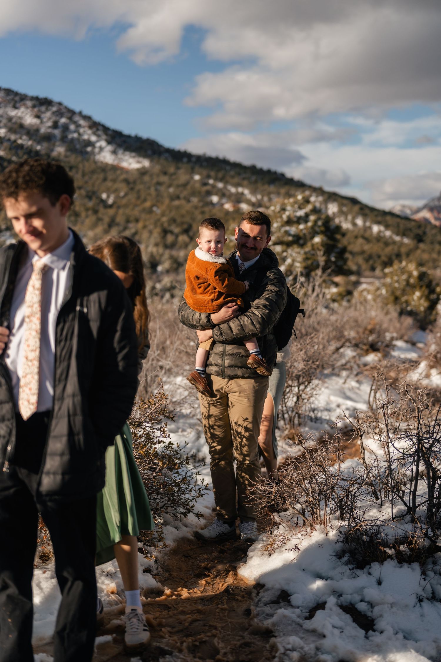 A winter wedding ceremony takes place on a snowy mountain trail with dramatic red rock cliffs in the background.