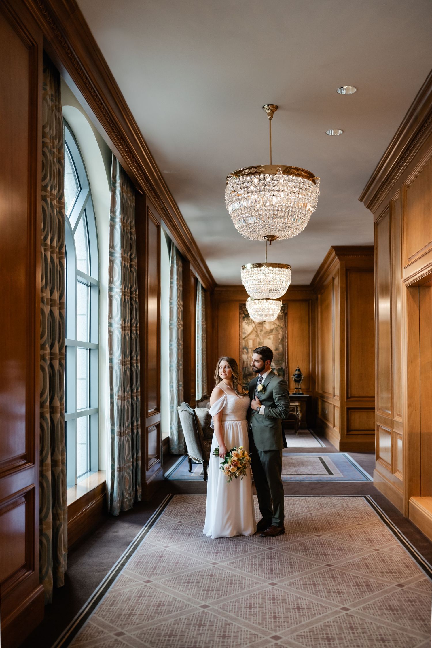 Elegant hallway with crystal chandeliers, wood paneling, and arched windows creates a luxurious wedding venue setting.