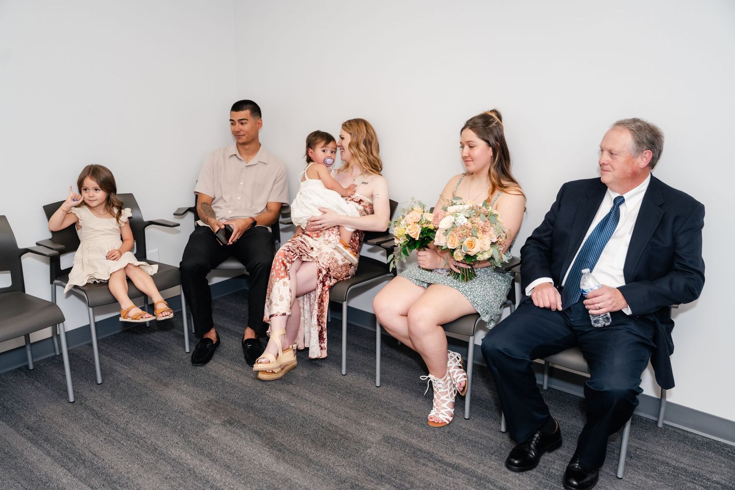 People sitting in chairs along a wall in what appears to be a waiting room or hallway area.