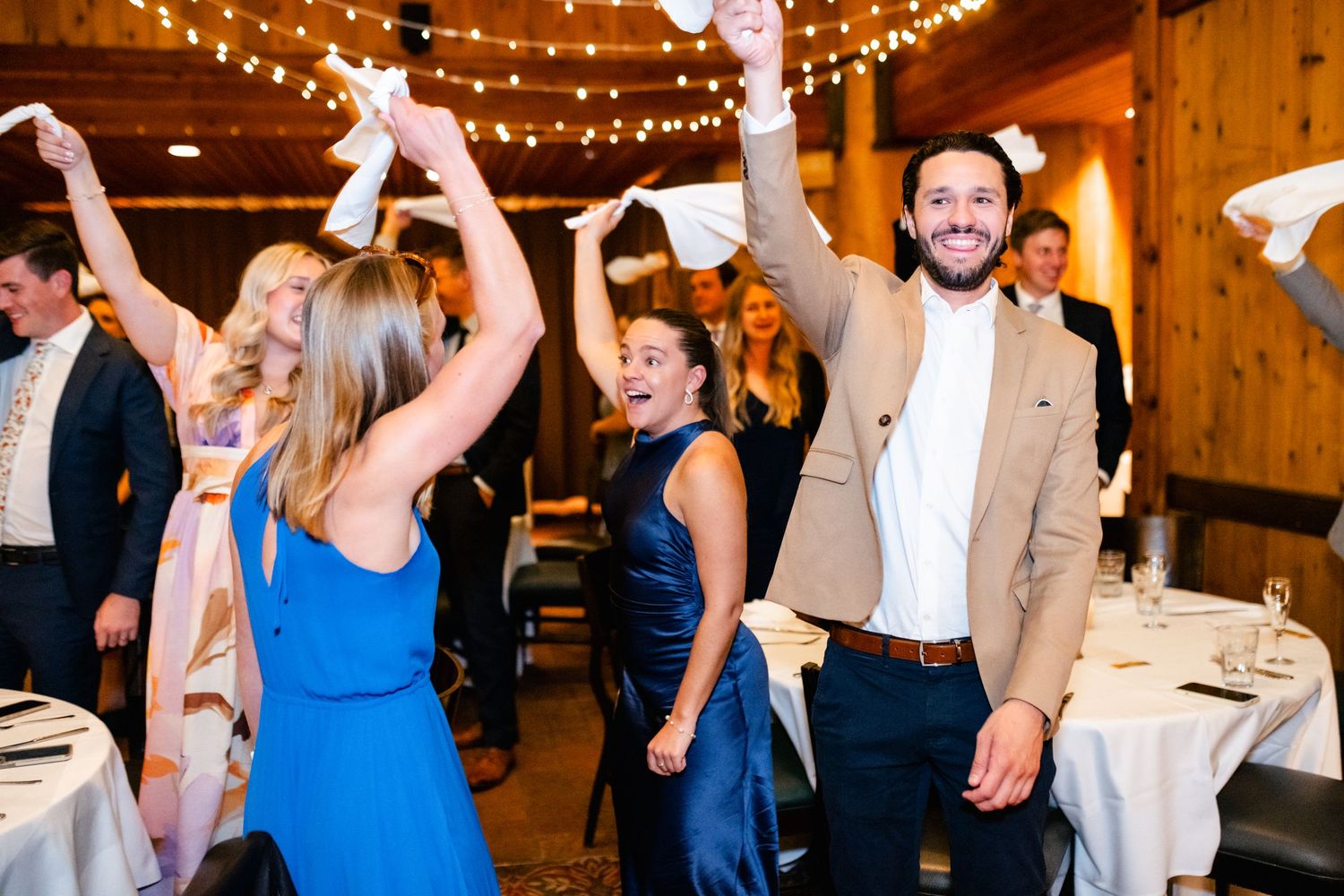 Wedding reception guests dancing and celebrating under warm string lights in a rustic wooden barn venue.