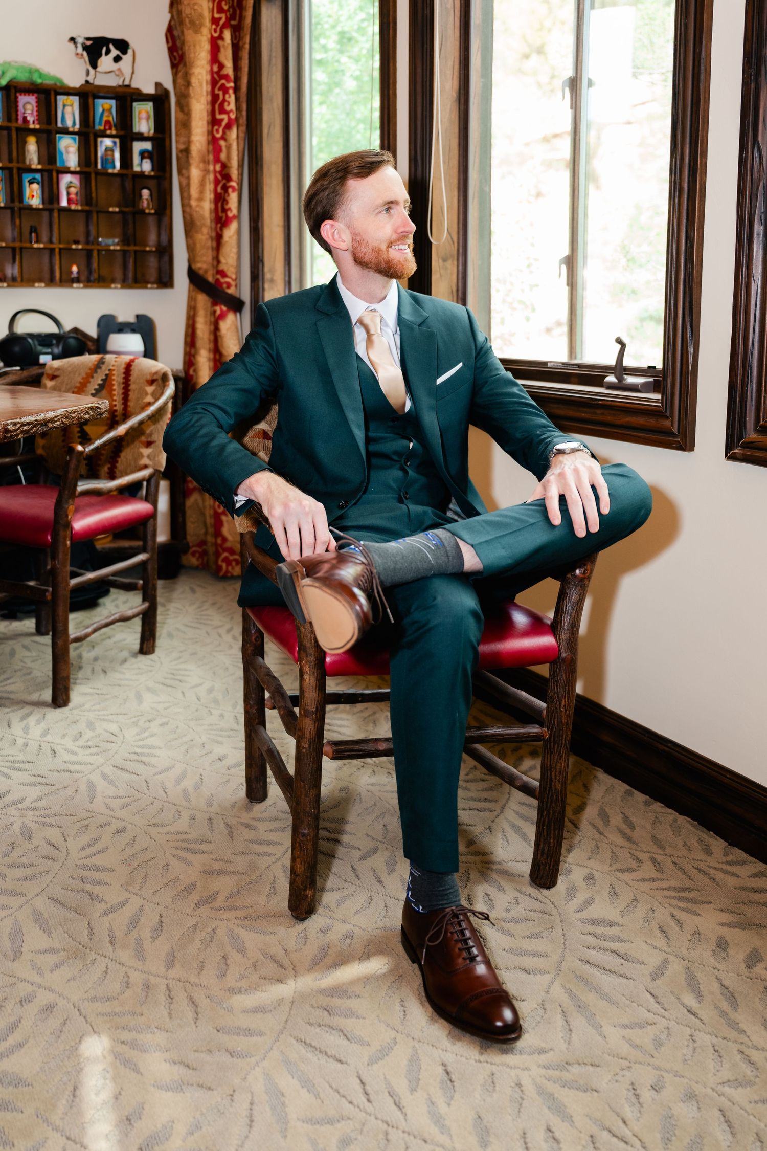 A person in a dark green three-piece suit and brown leather shoes sits in a wooden chair by a window.