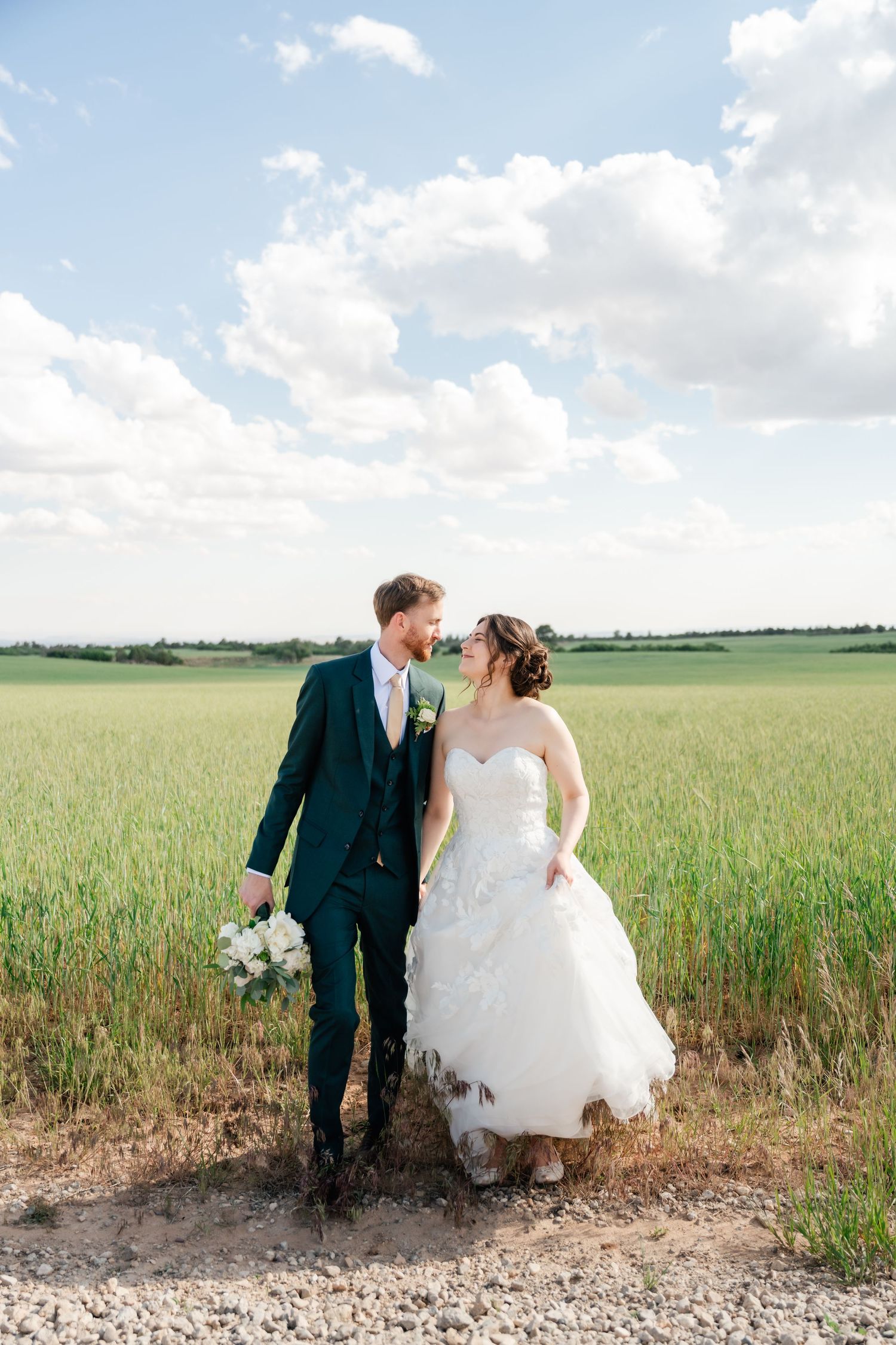 A romantic wedding couple sits together at the edge of a lush green wheat field on a bright summer day.