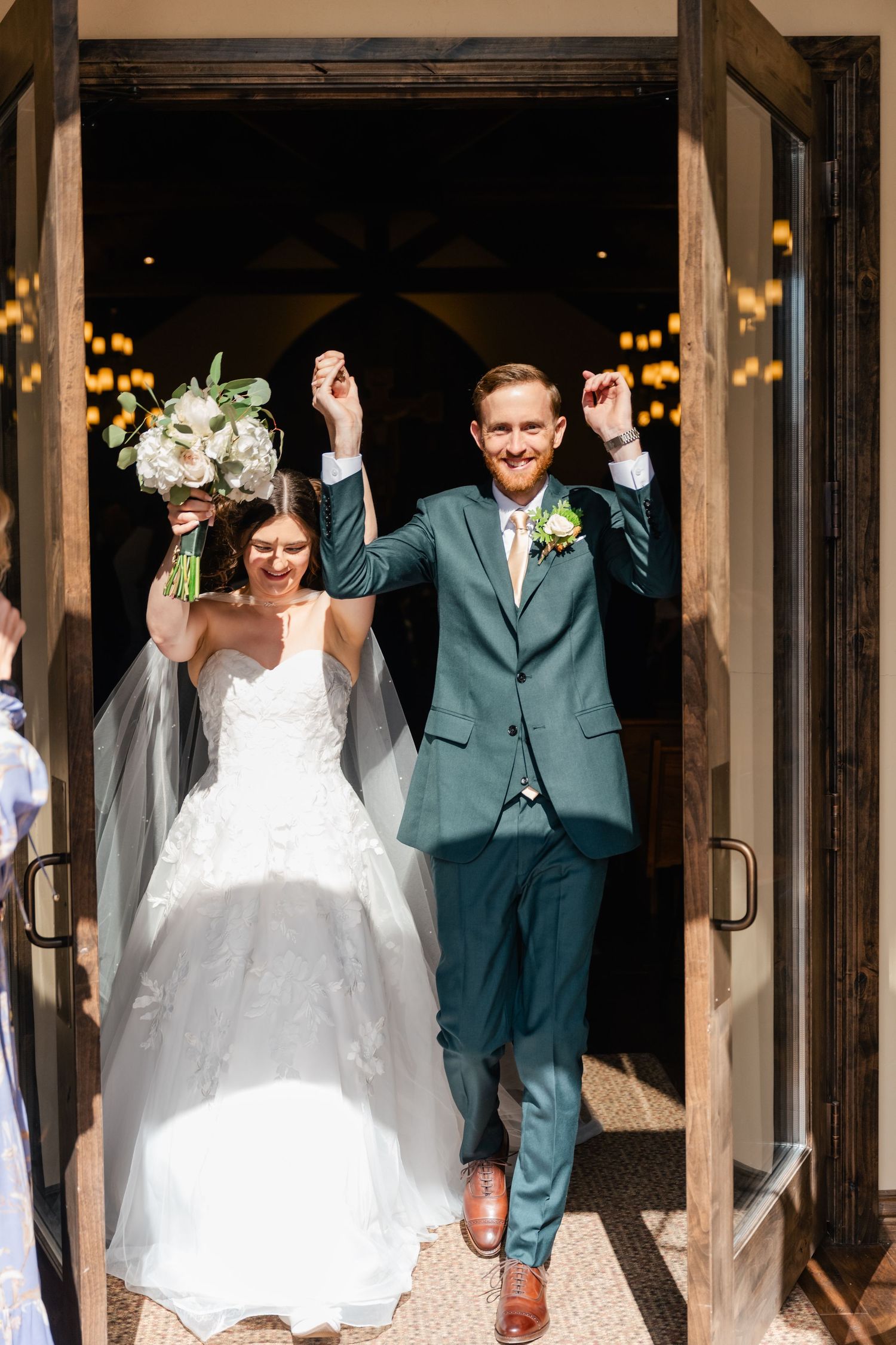 Newlyweds celebrate with raised arms as they exit their wedding ceremony through wooden doors.