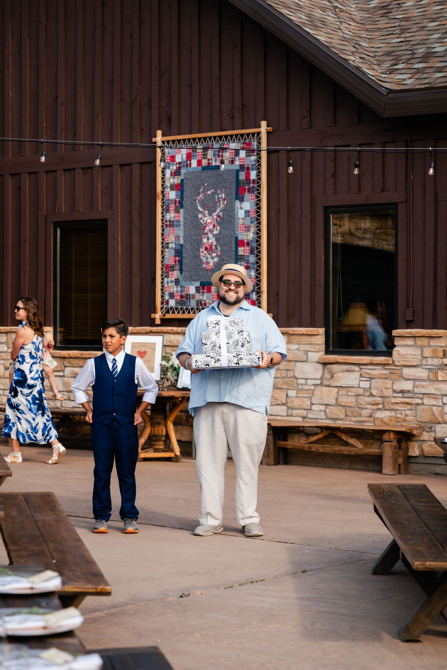 Wedding guests gather around wooden picnic tables outside a rustic stone and wood lodge with decorative quilts on display.