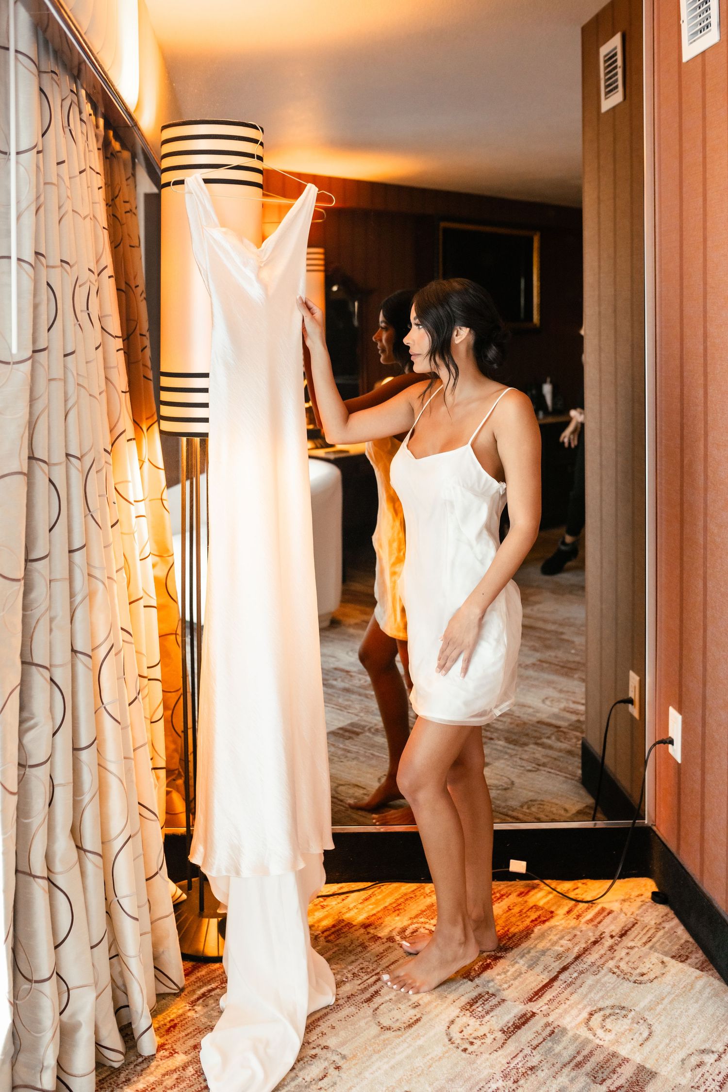 Couple in white getting ready in a warmly lit hotel hallway with patterned carpet and curtains.