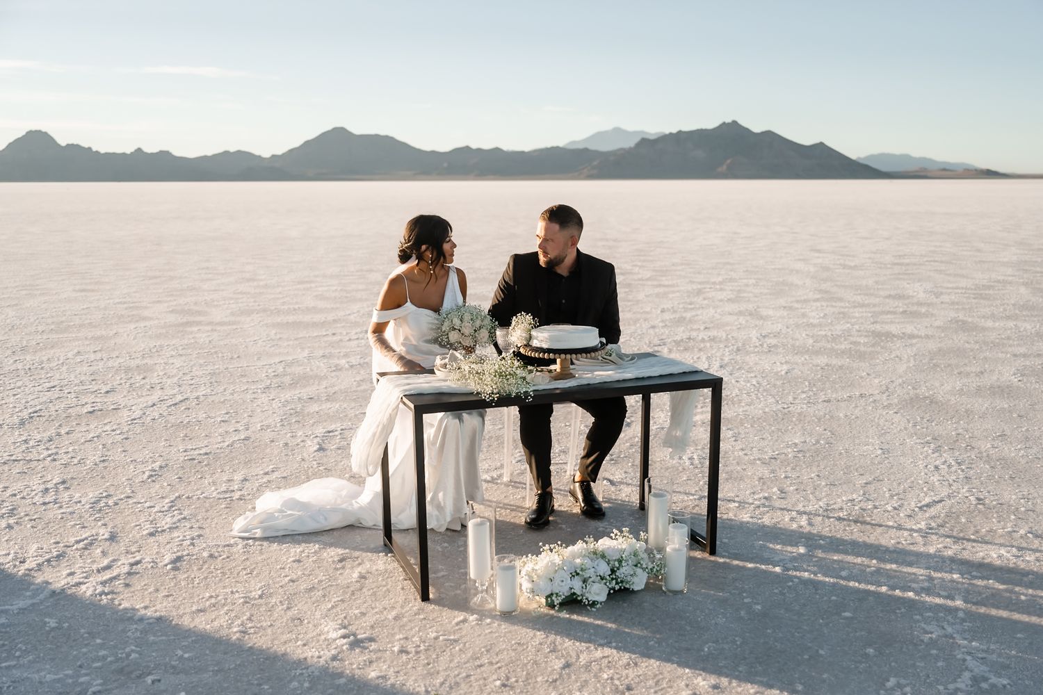A couple sits at a table on the pristine white salt flats with mountains visible in the distance.
