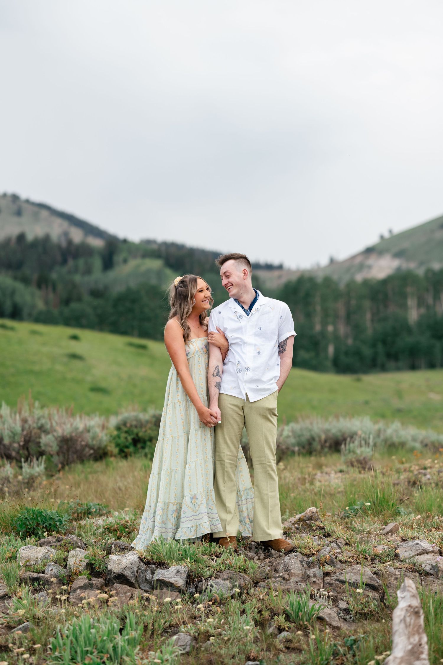 A couple stands together on a rocky hillside surrounded by green mountains and meadows in a romantic meadow. Utah Engagement Photographer.