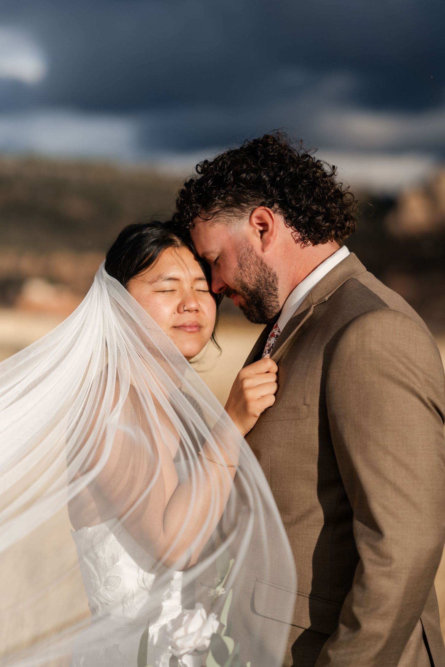 A romantic wedding couple shares intimate moments under a flowing veil against a mountain backdrop.
