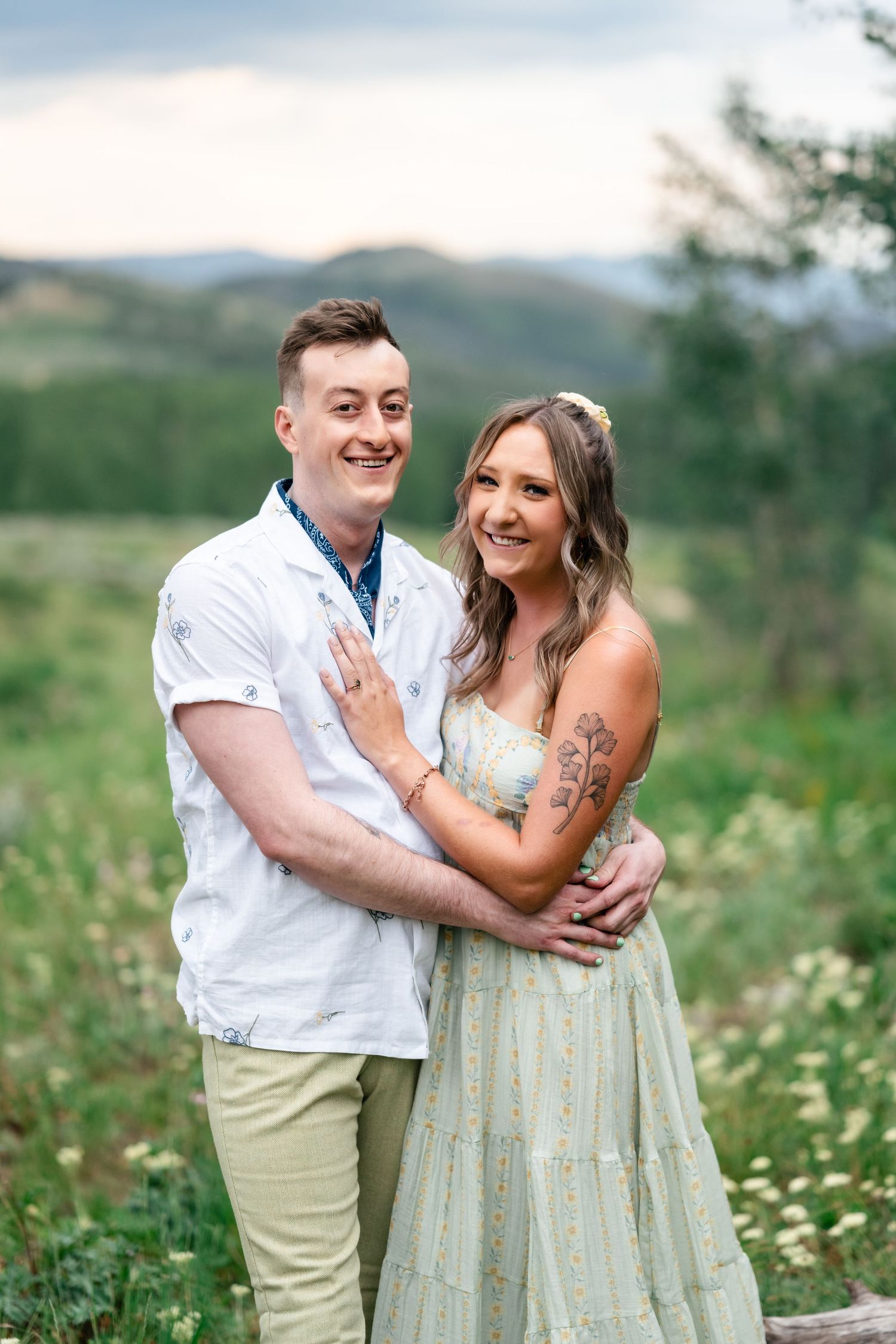 A couple shares joyful laughter together in a scenic meadow with wildflowers and mountains in the background.