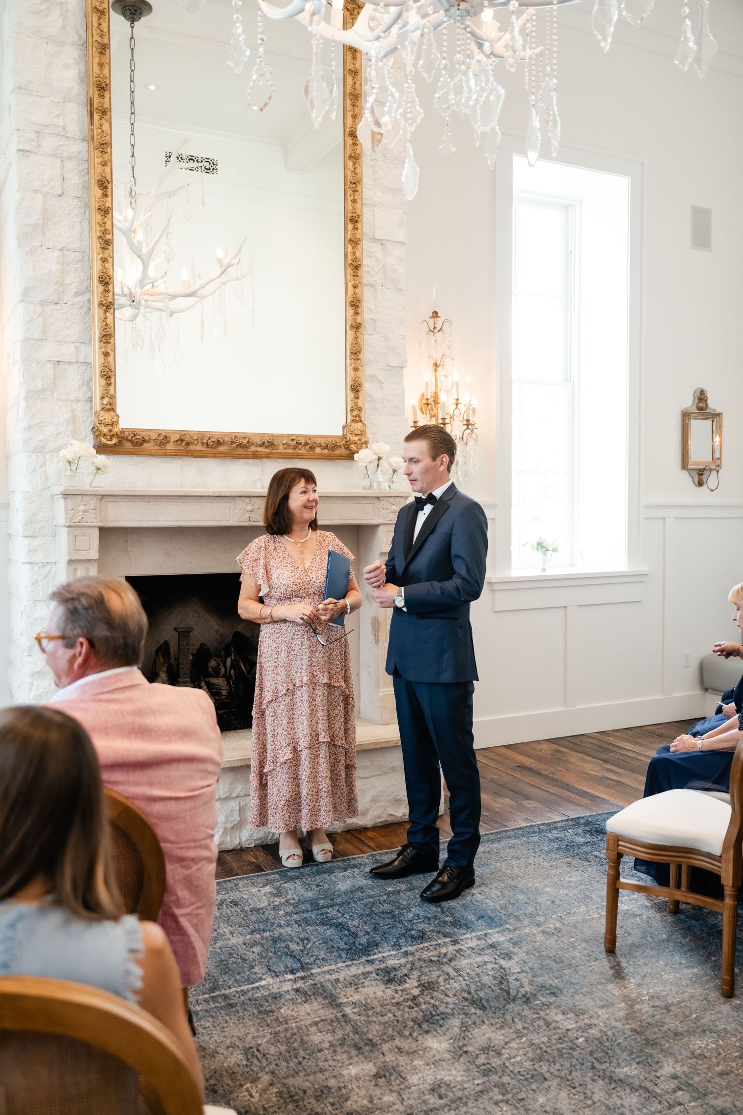 An elegant wedding ceremony in a white room with ornate gold-framed mirror and chandelier.