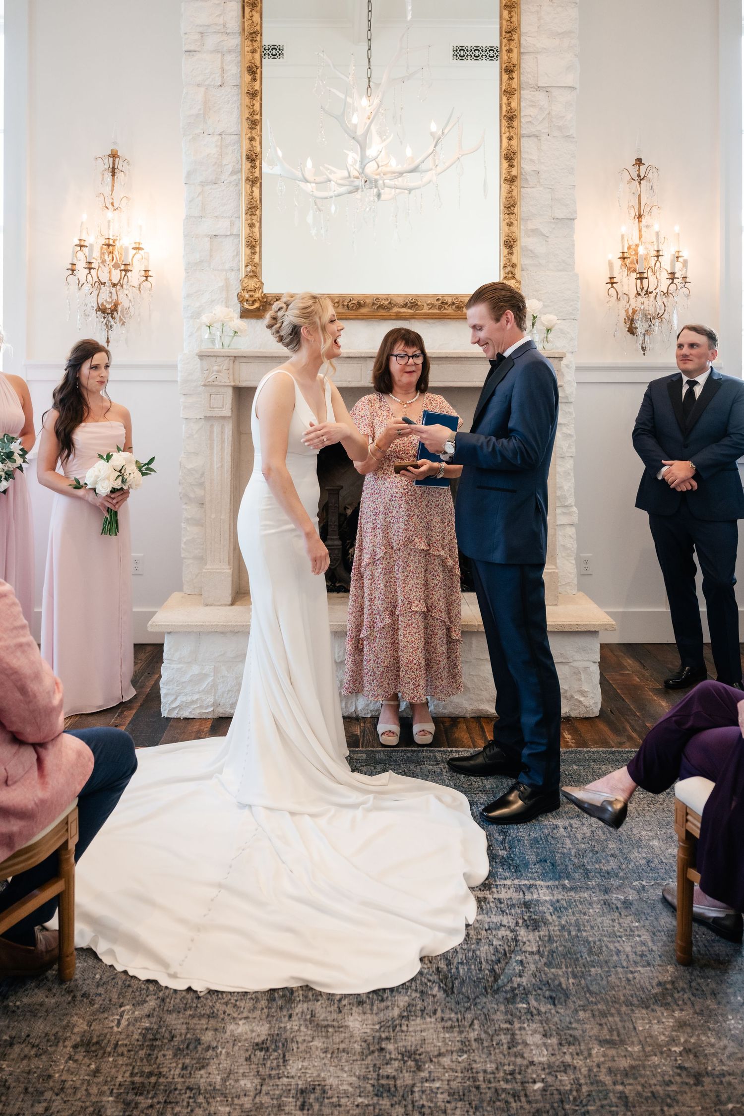 A wedding ceremony takes place in an elegant room with white walls and crystal chandeliers.