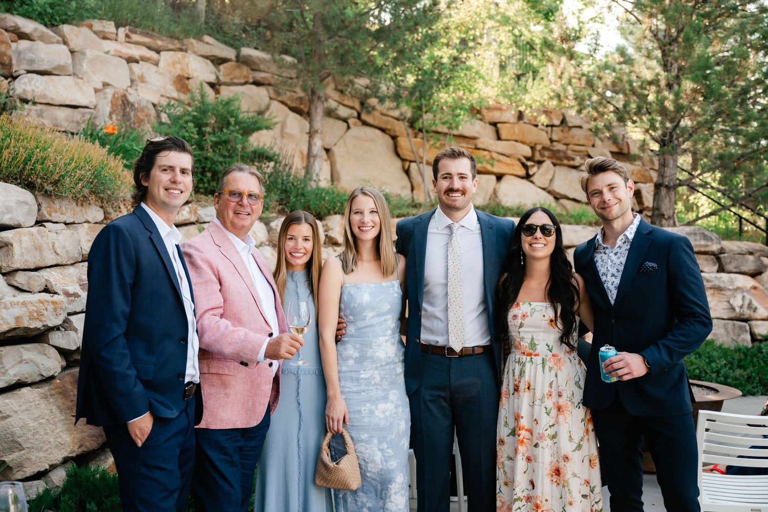 Wedding party members pose for photos against a rustic stone wall in an outdoor setting.