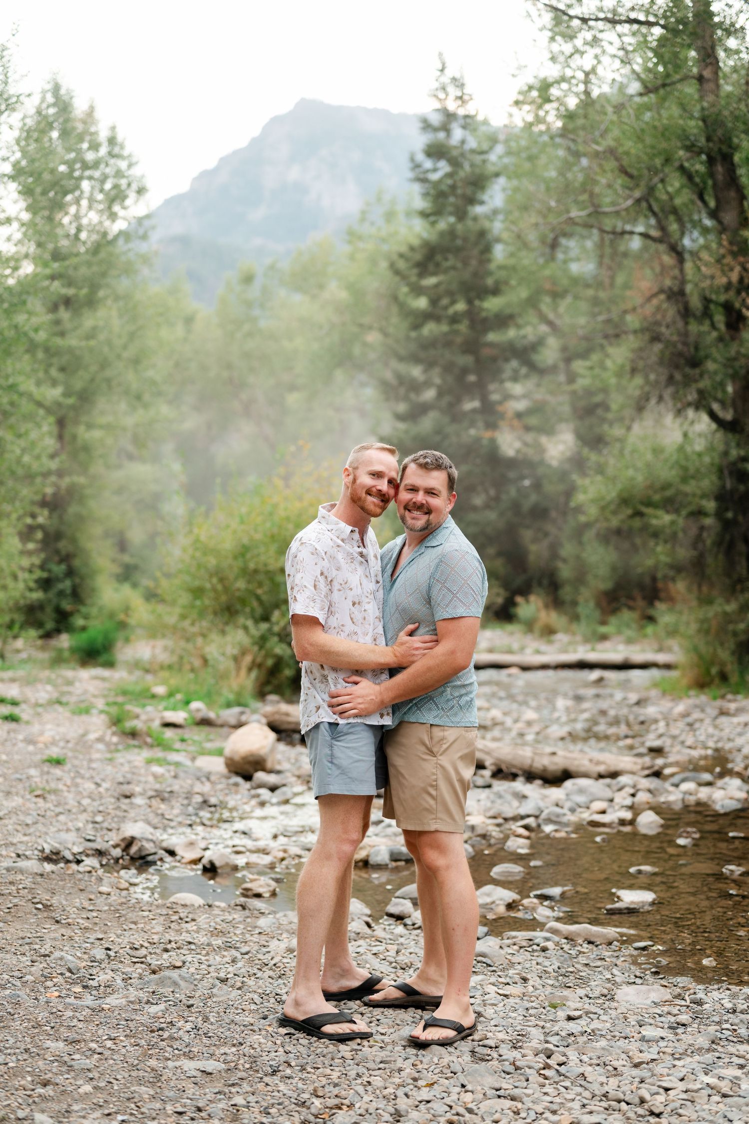 Two men embracing and smiling on a rocky riverbed with mountains and evergreen trees in the background.