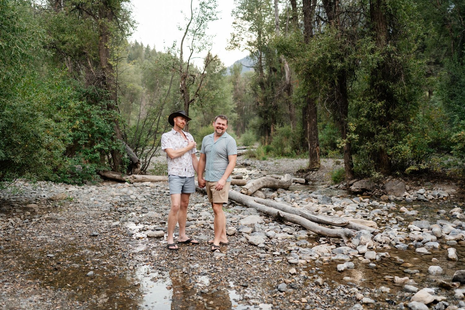 LGBTQ Couple standing together on rocky riverbank surrounded by lush green forest trees in a natural outdoor setting during engagements.