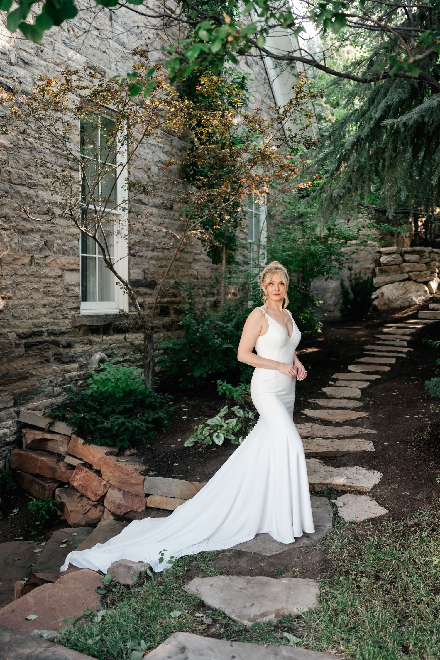 Bride poses in fitted mermaid wedding gown with long train beside rustic stone building and garden path of the Washington School House Hotel