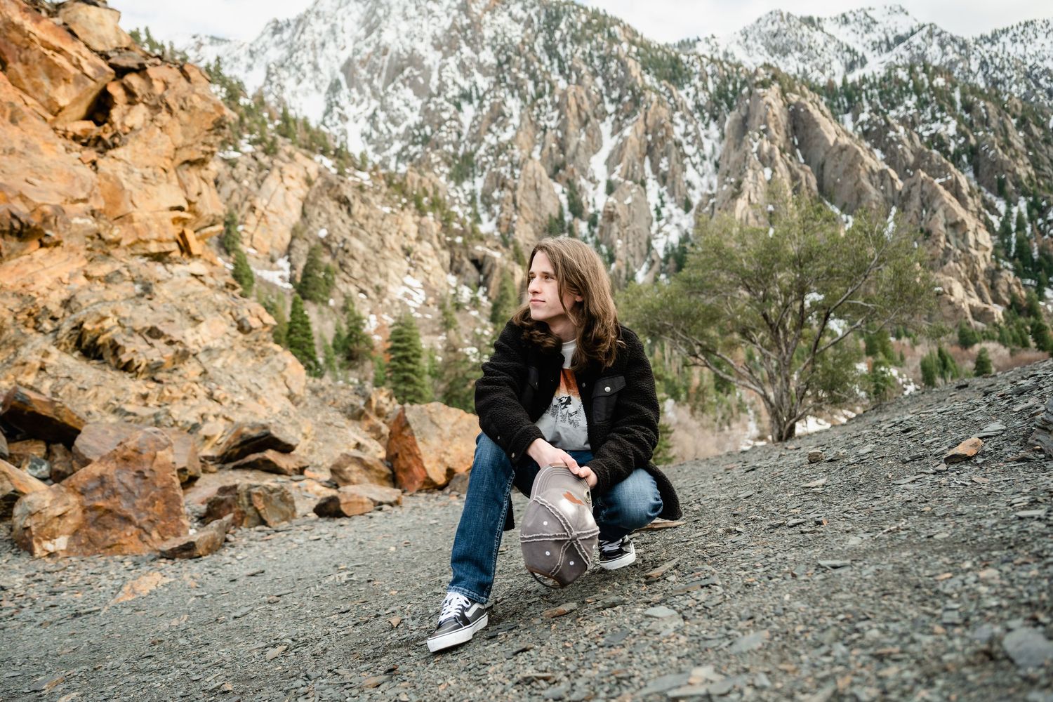 A person crouches on a mountain trail with snow-capped peaks and orange rocky cliffs in the background.