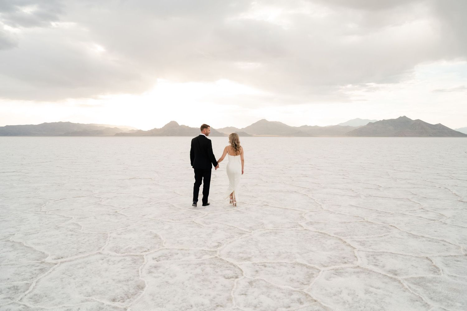 Salt Flats Elopement Photographer. A couple stands together on the vast white salt flats with mountains and cloudy skies in the distance.