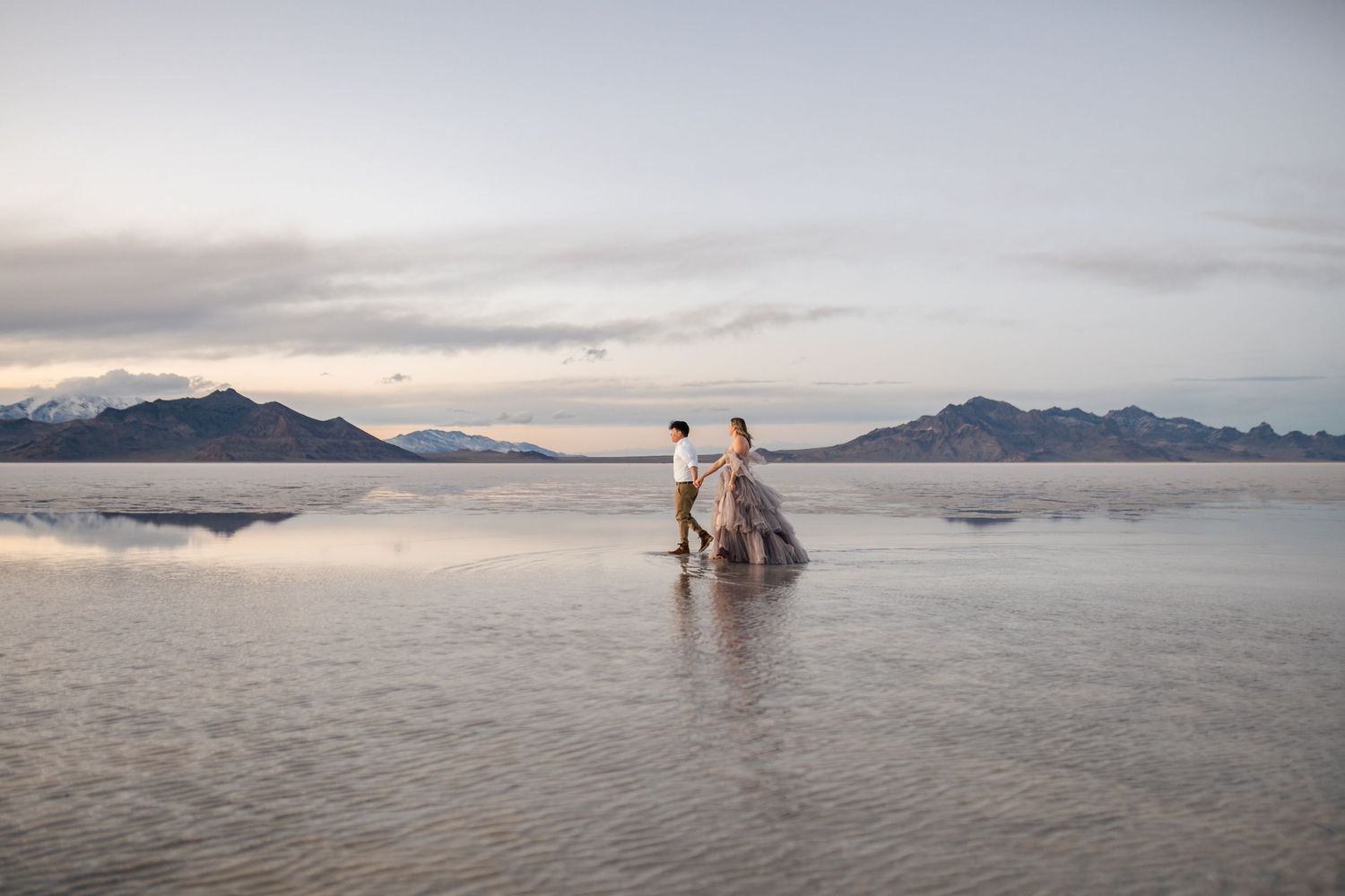 Utah Salt Flats Photographer. Silhouettes stand in shallow reflective water against a pastel sunset sky with mountain ranges on the horizon.