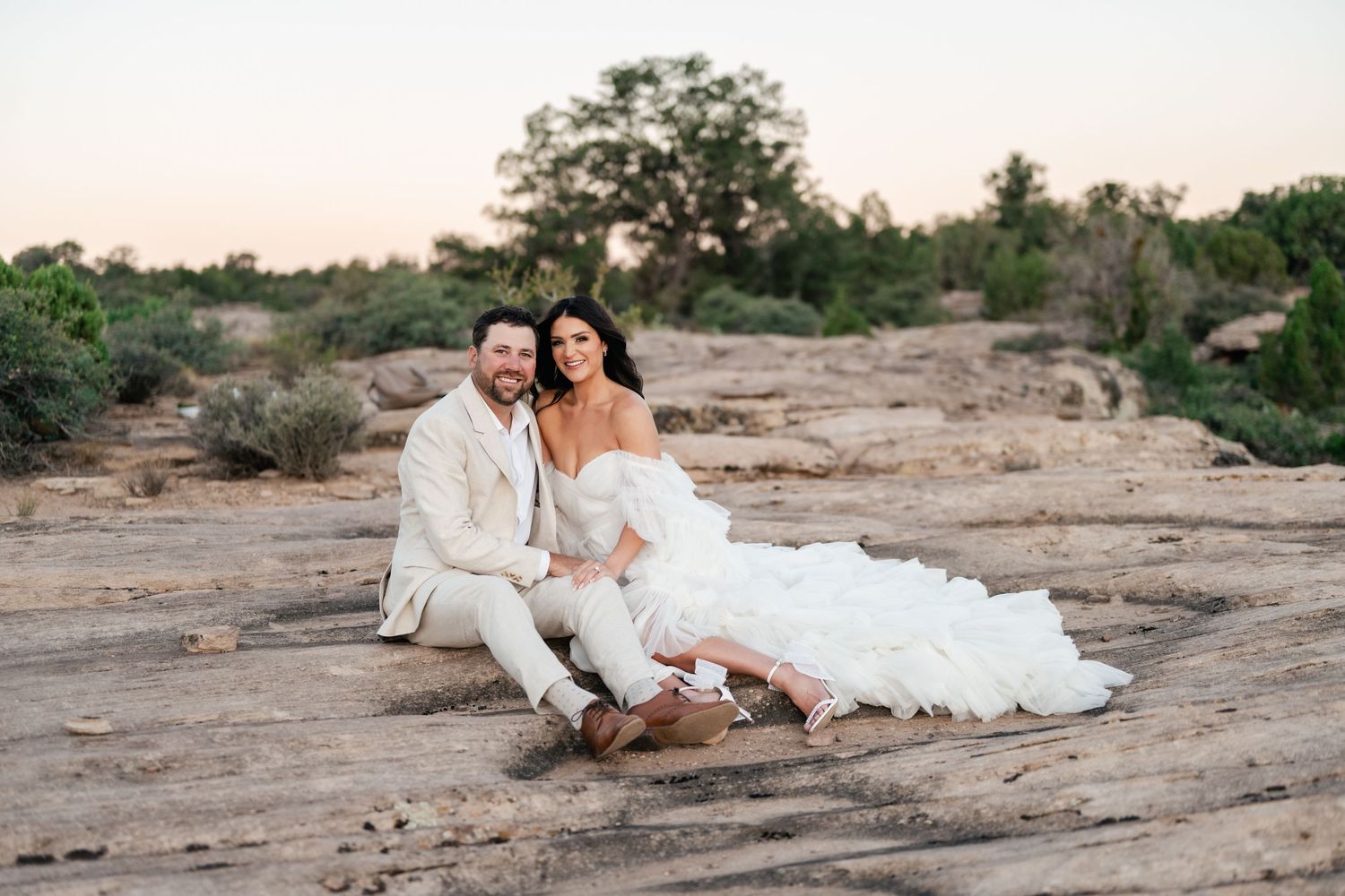 Zion destination photographer. A couple in wedding attire sits together on red desert rocks during sunset in a romantic outdoor setting.
