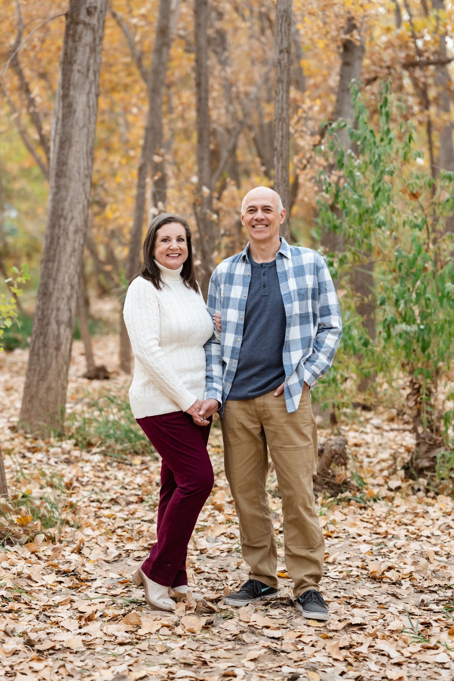 A couple poses together on fallen autumn leaves in a wooded setting wearing coordinated casual attire.