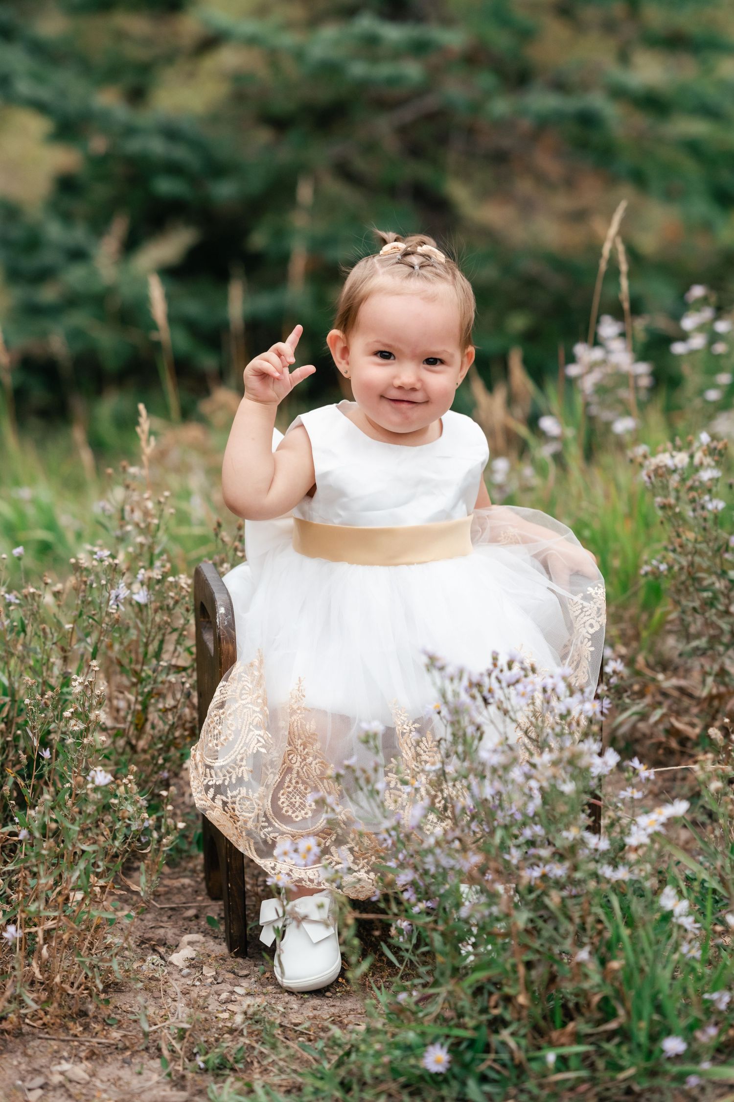 A small child in a white dress sits among wildflowers in a natural outdoor setting.