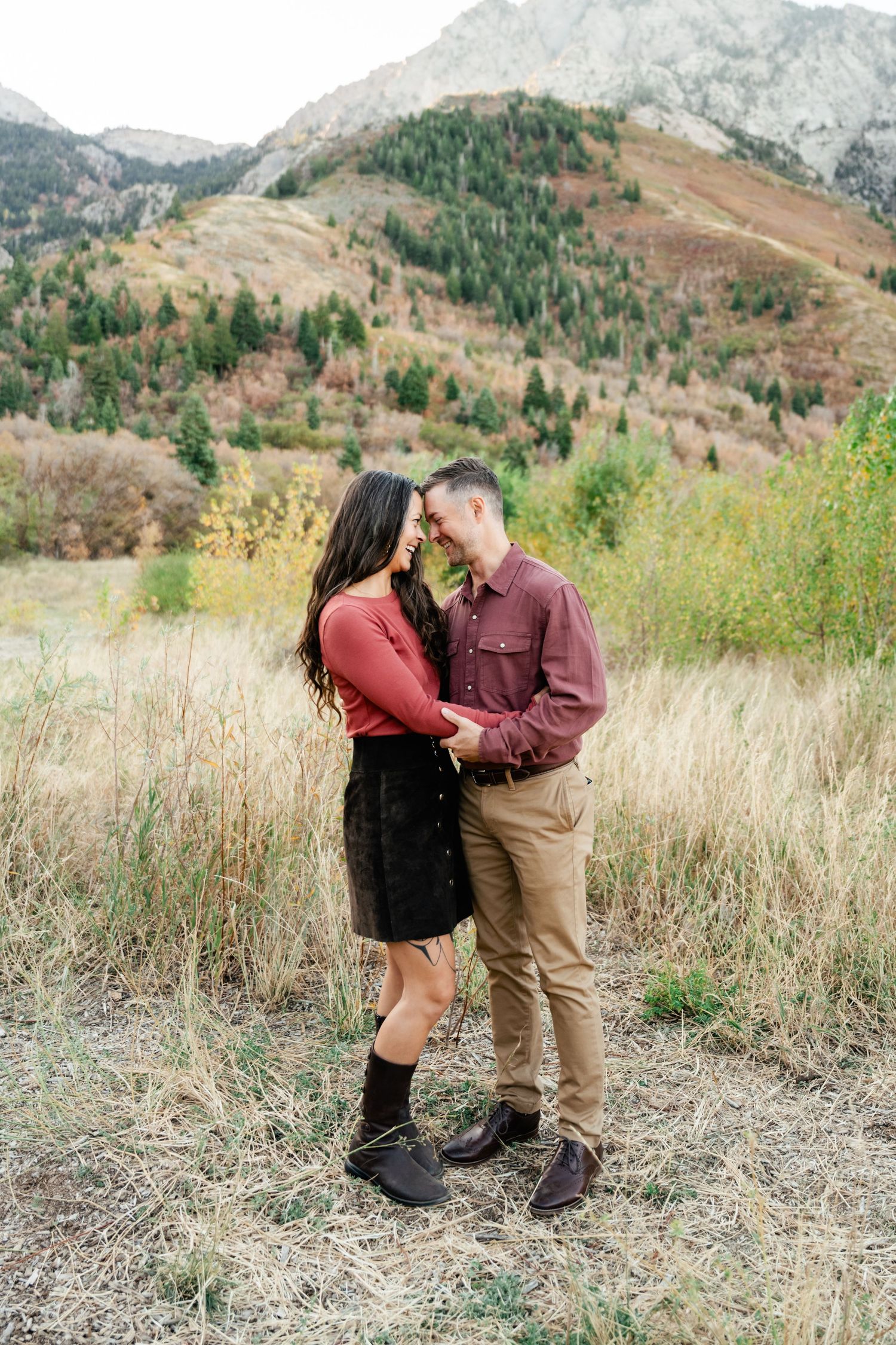 A couple shares an intimate moment in tall grass with majestic mountain scenery behind them.