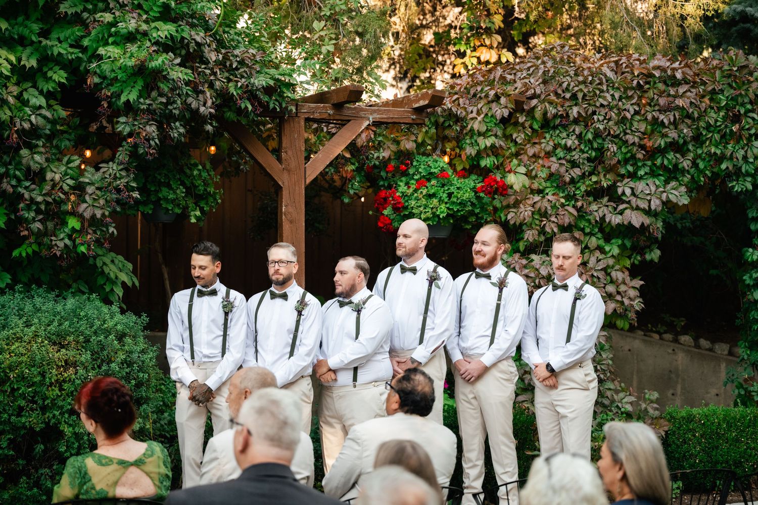 Wedding party groomsmen wearing white pants with suspenders standing in formation during outdoor ceremony.