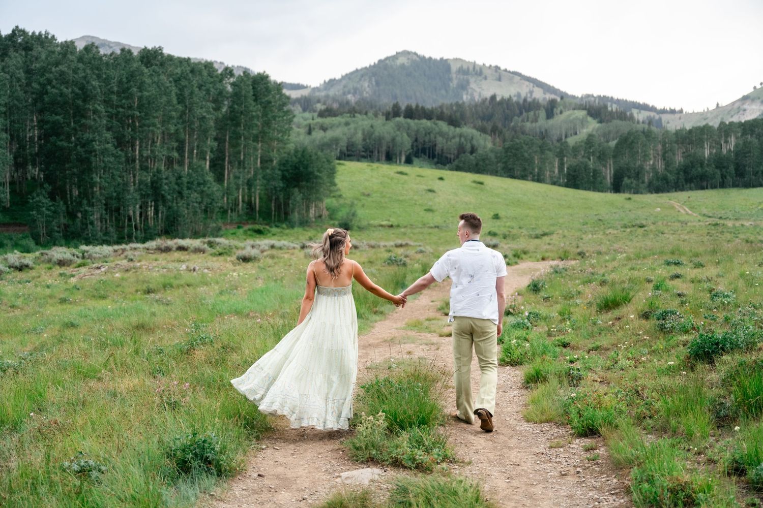 Two people walking hand in hand along a mountain trail surrounded by evergreen forest and meadows in a scenic landscape. Utah Wedding Photo