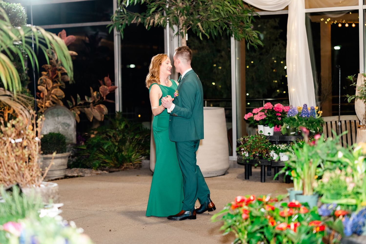Two people in elegant matching emerald green formal attire dance on an outdoor patio surrounded by tropical plants and flowers.