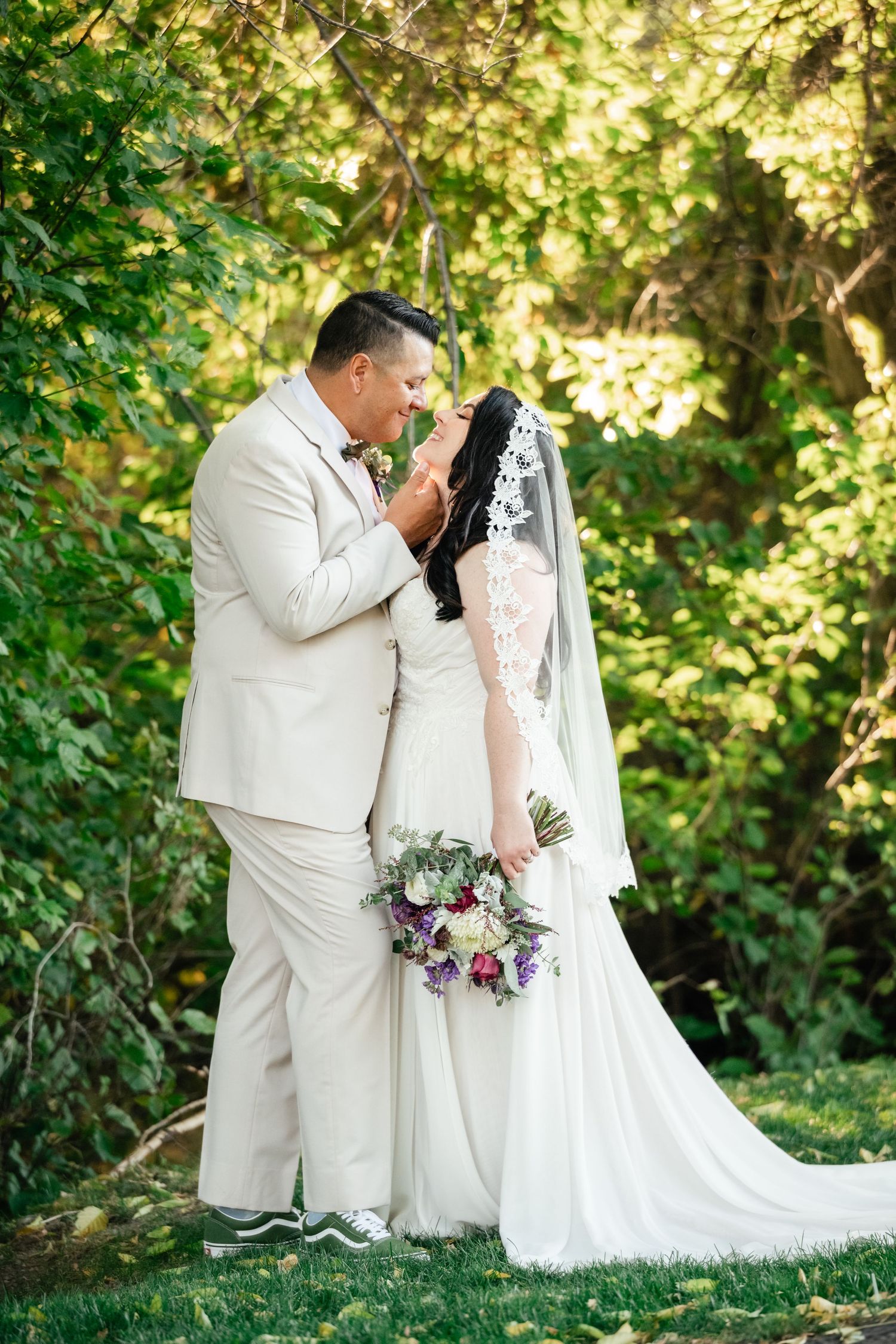 A newlywed couple poses in a garden setting with the bride holding a purple and white floral bouquet.