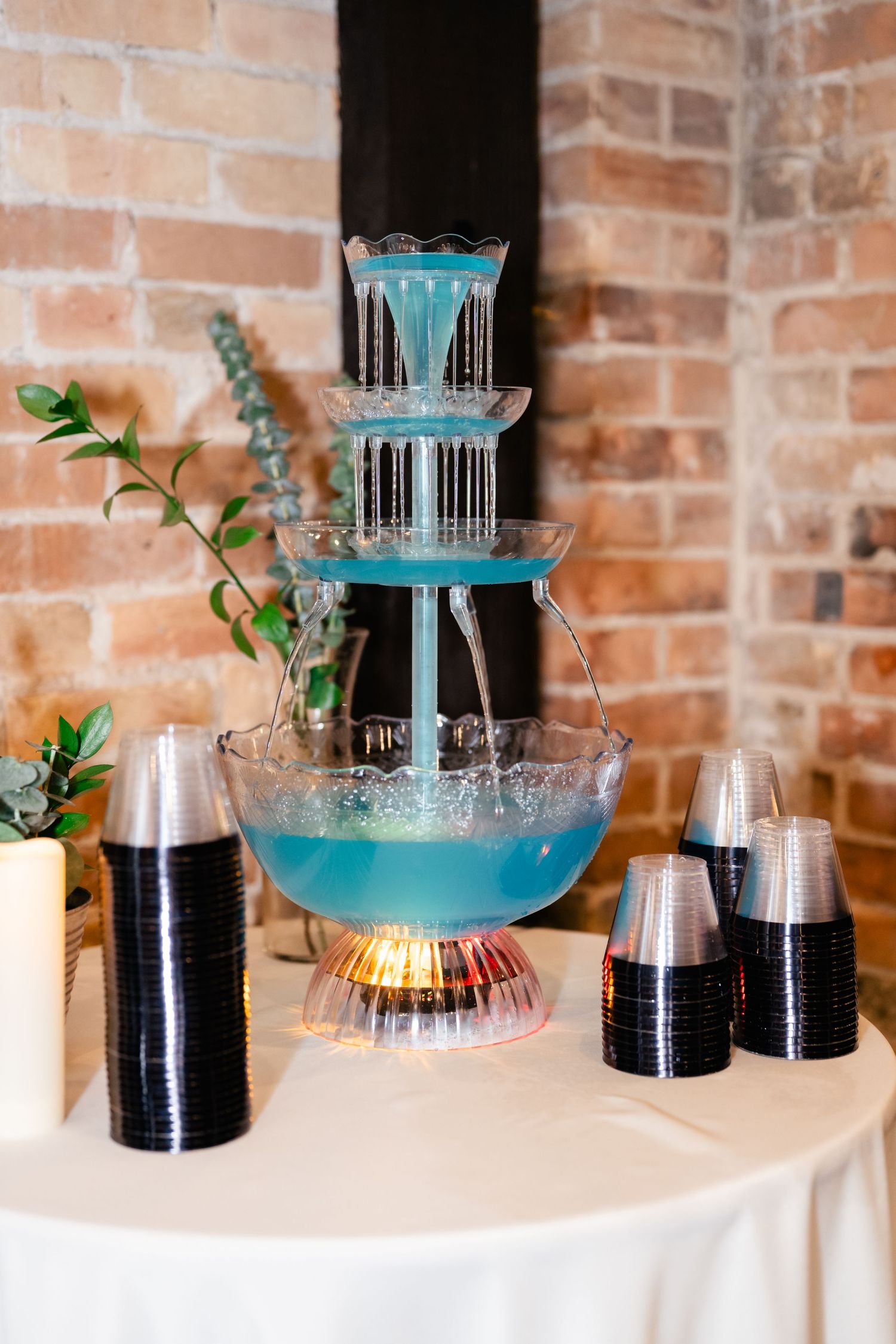 Elegant blue cocktail fountain with flowing tiers sits on a white tablecloth with black plastic cups and greenery accents.