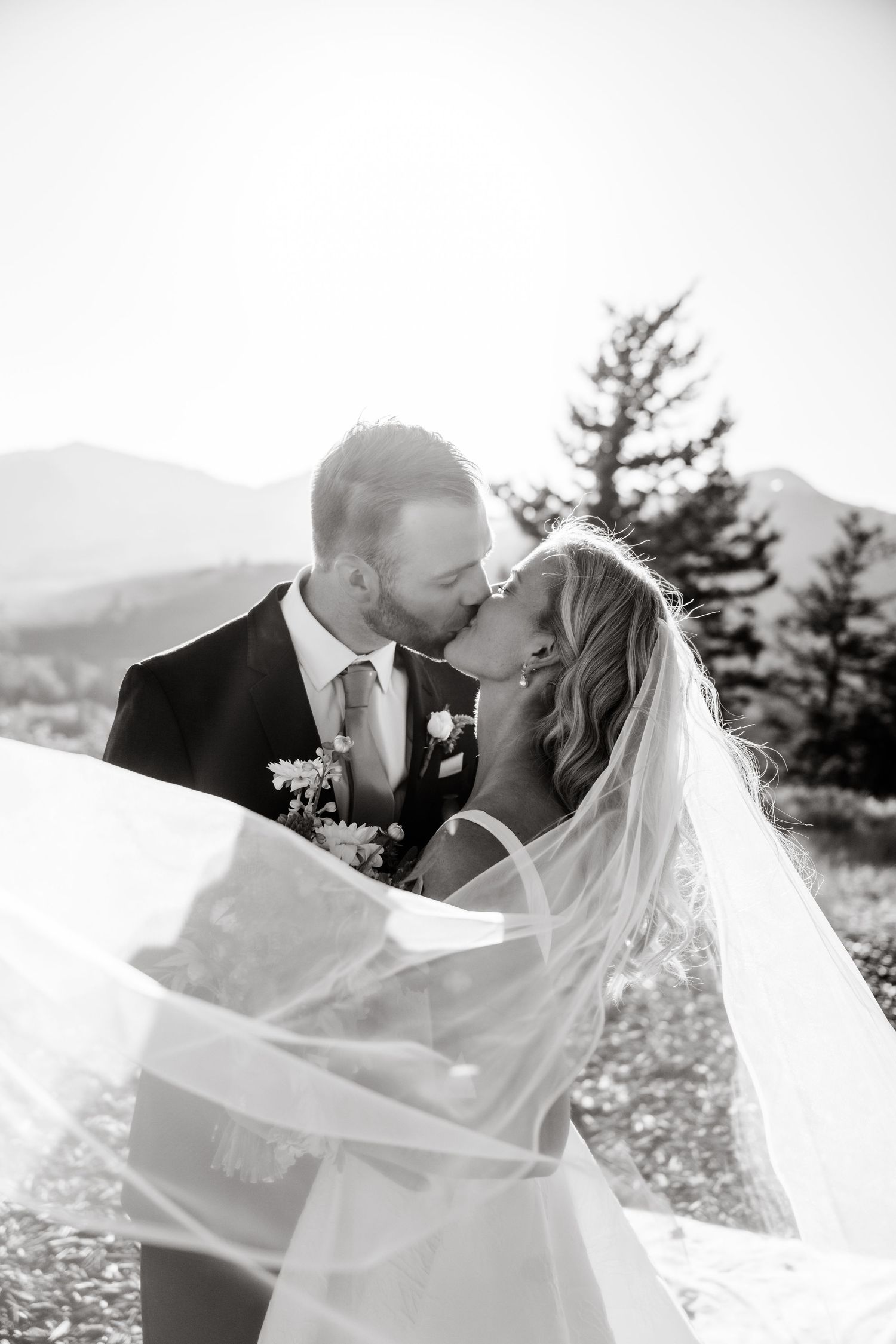 Romantic black and white wedding photo with couple kissing on scenic mountaintop as bride's veil flows in the wind. Park City Wedding Photo.