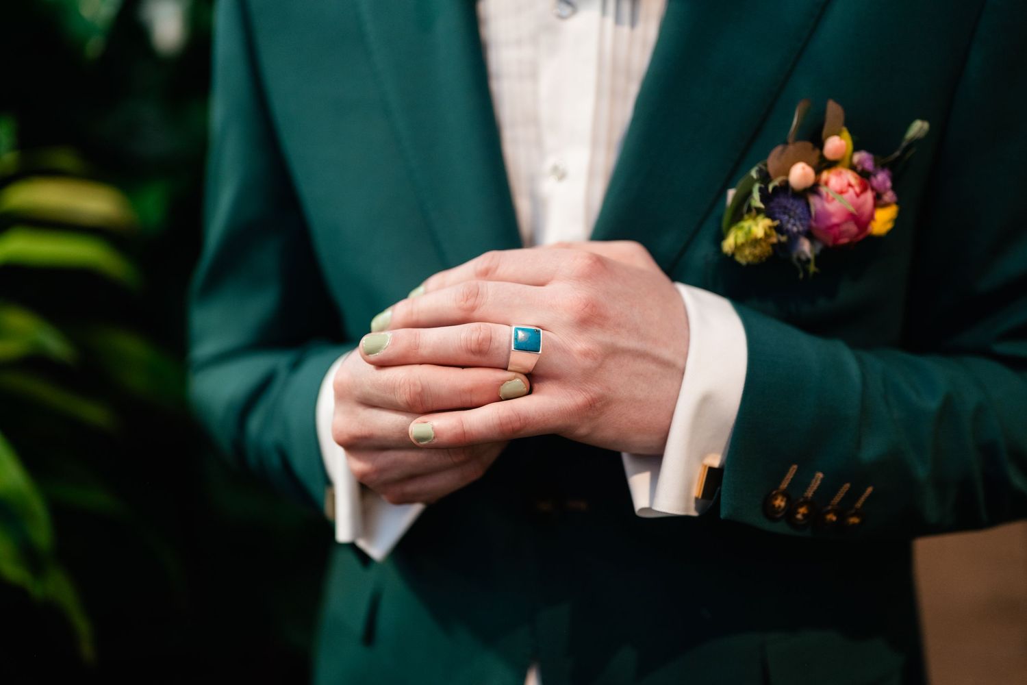 Close-up detail of hands wearing an emerald green suit jacket and wedding ring with boutonniere in background.