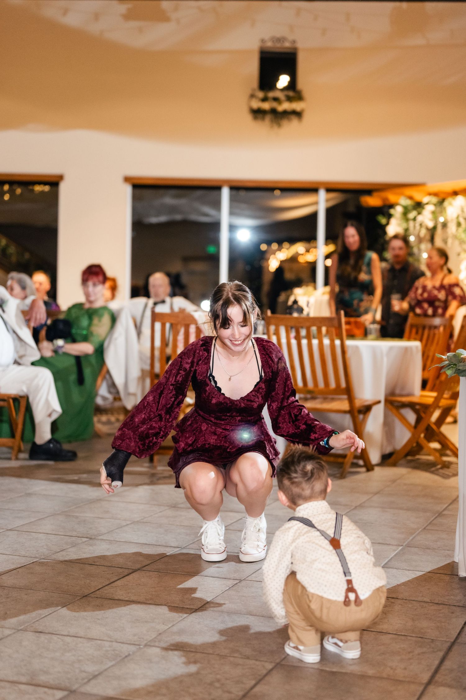 A woman in a burgundy dress crouches down to dance with a toddler in suspenders on a tiled floor at a reception.