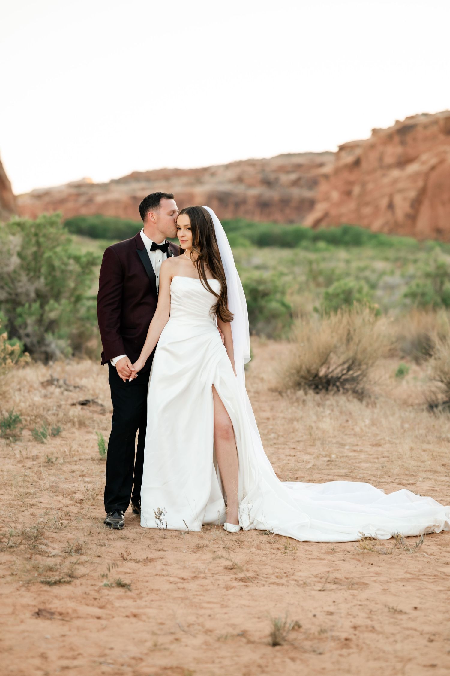 Bride and groom embrace in desert landscape with red rock formations, she wears a white gown with veil, he wears burgundy tuxedo.