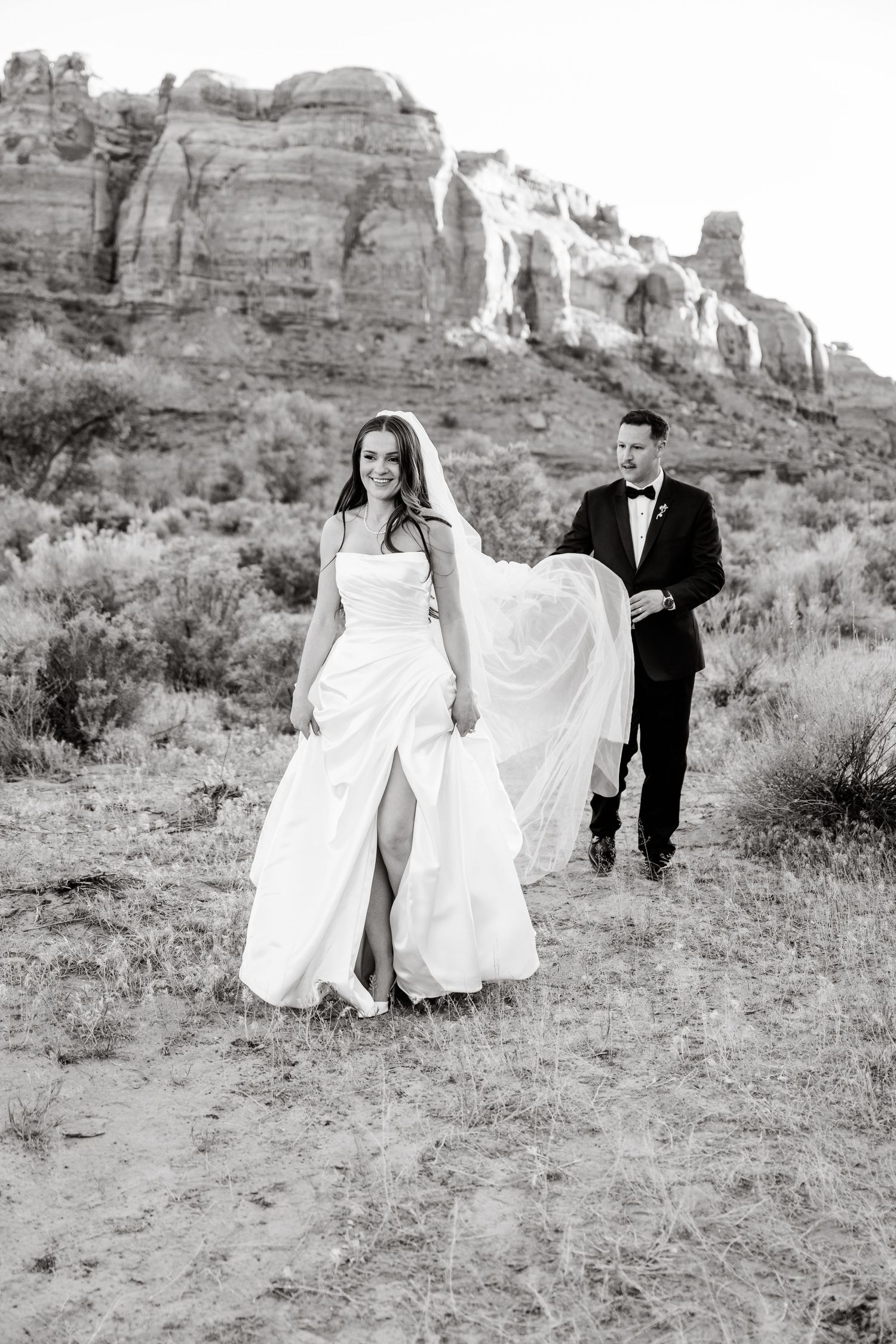 Black and white photo of smiling bride in flowing gown walking ahead of groom through desert canyon terrain.