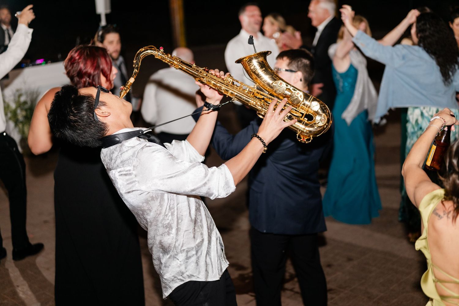 Two saxophonists perform energetically on a wedding dance floor at night, entertaining guests at a lively reception.