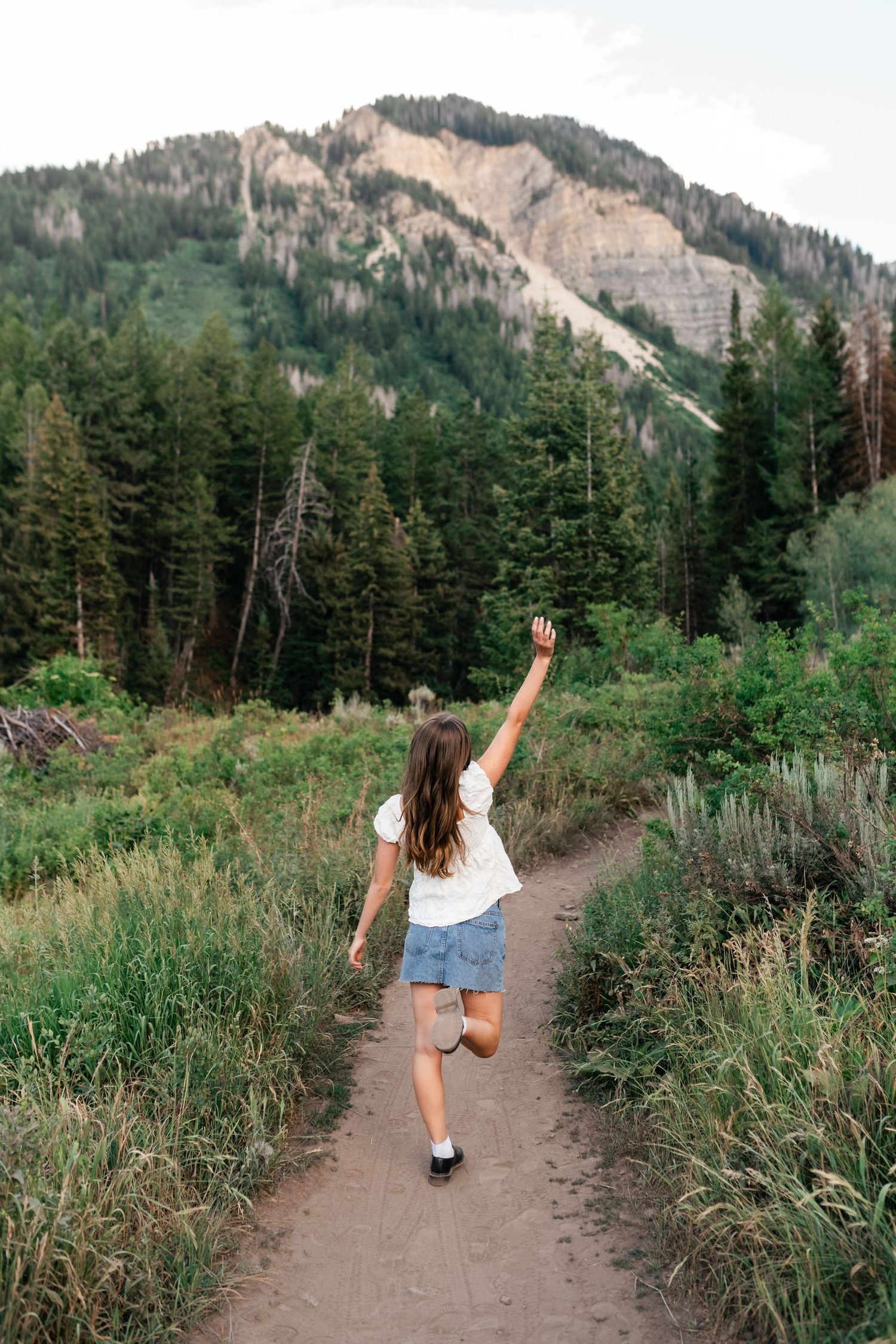 People walk and pose playfully on a dirt hiking trail surrounded by evergreen trees and mountain views.