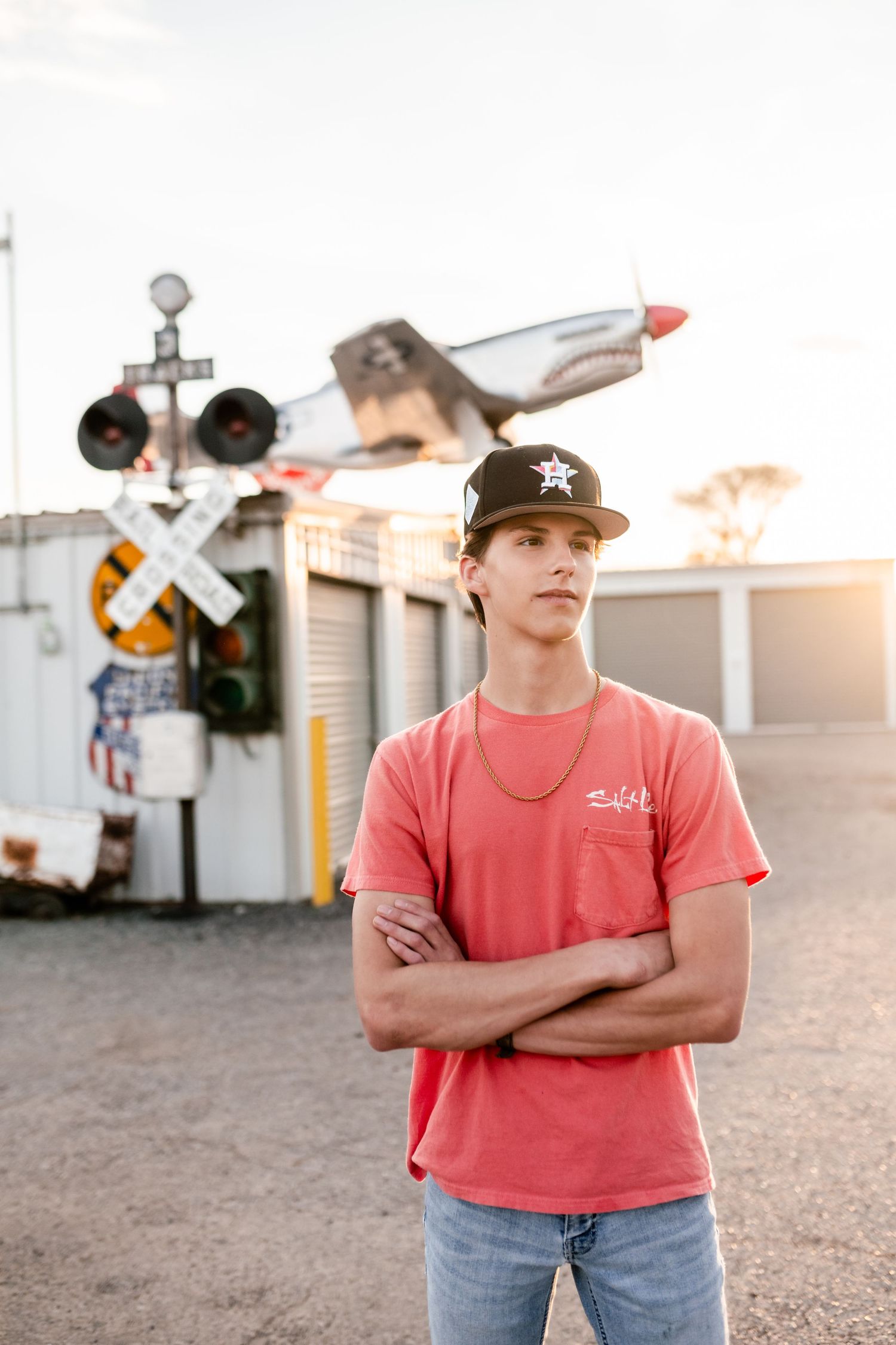 Person in a red t-shirt and baseball cap stands in front of a railroad crossing with a vintage aircraft display at sunset.