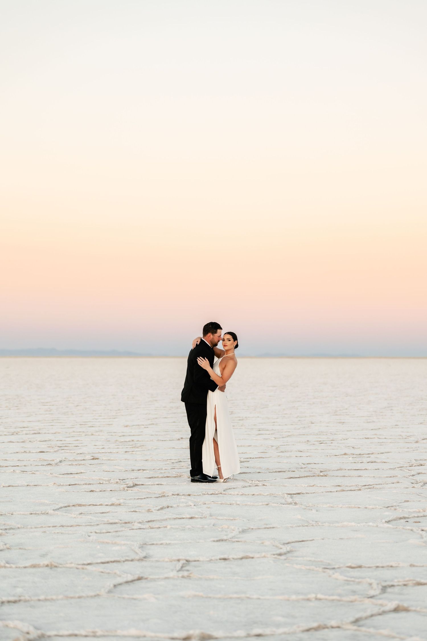 Utah Salt Flats Photographer. A wedding couple embraces on salt flats during a pastel sunset.