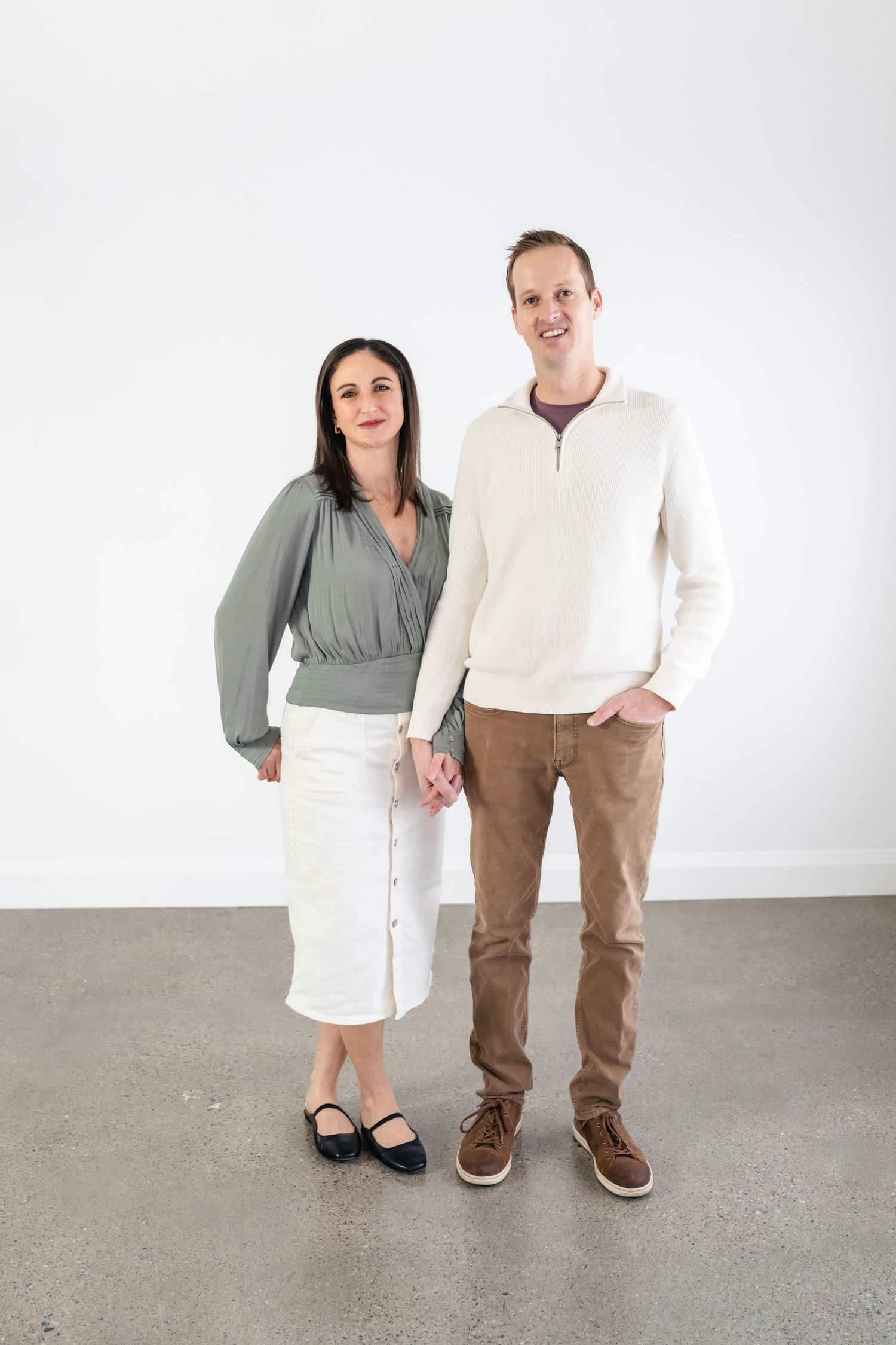 A couple stands together against a white wall wearing casual clothing and holding hands.
