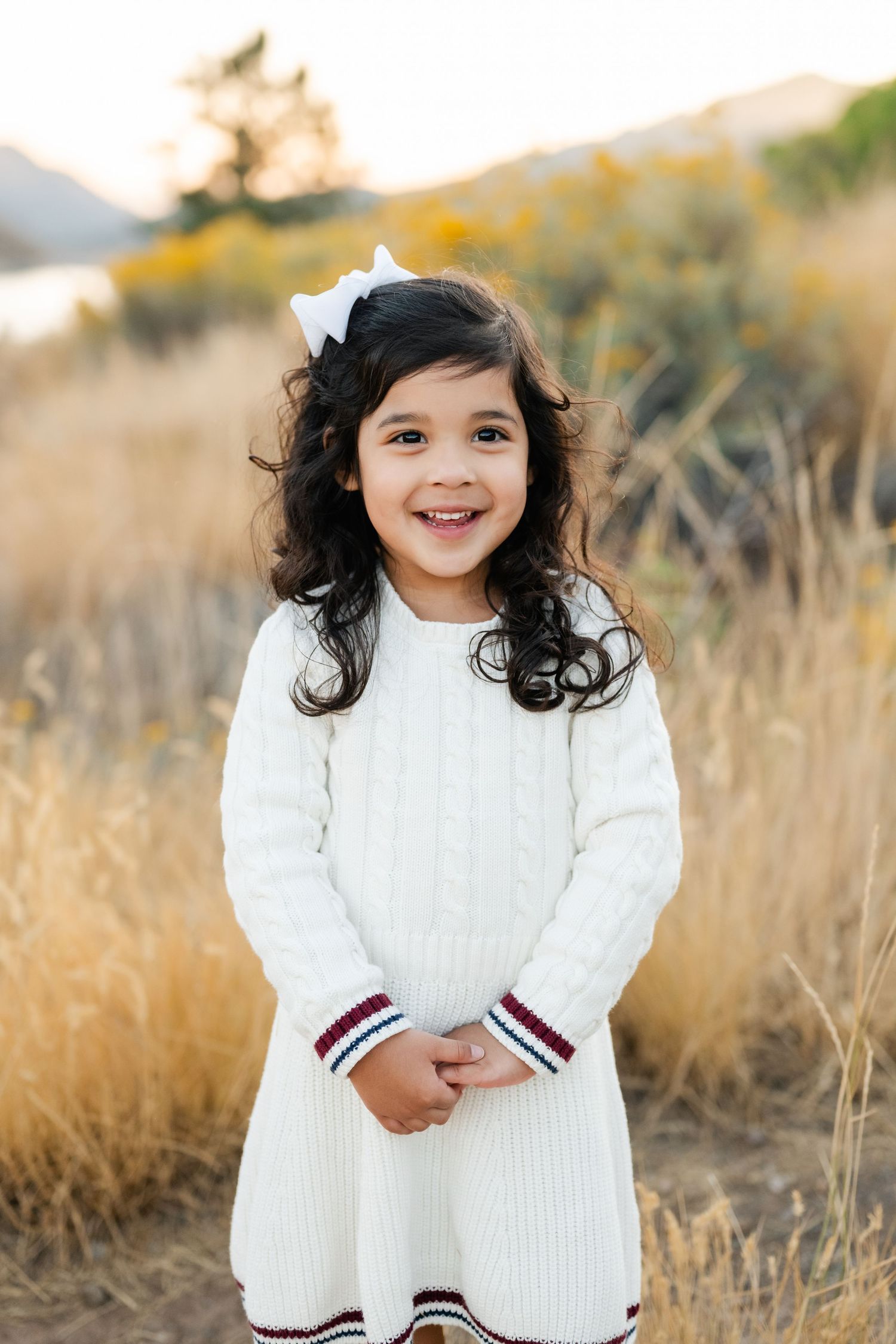 A child wearing a white knit dress with striped trim stands in a golden field during sunset.